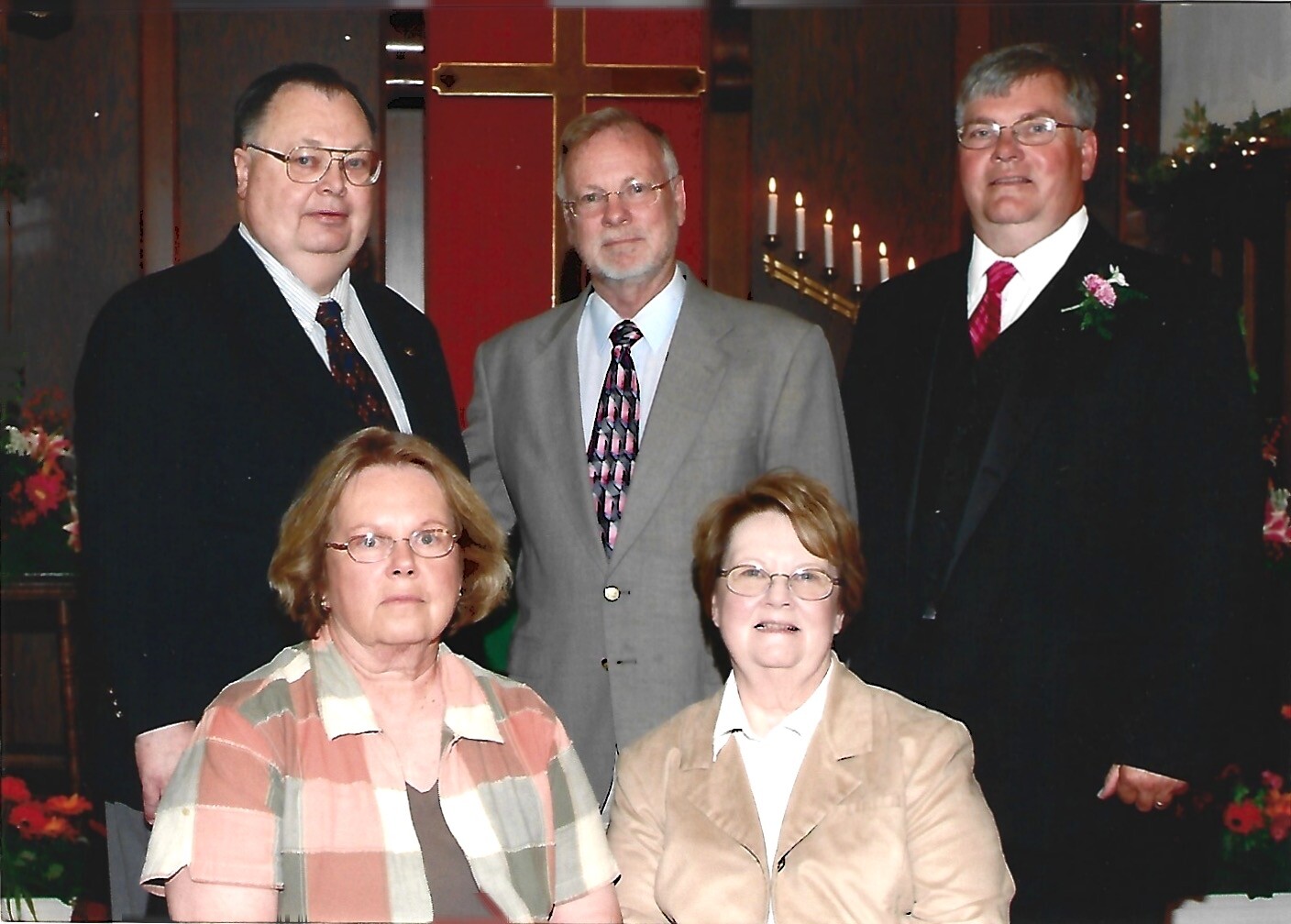 Five adults in formal attire pose for a group photo in a church, with a cross and lit candles in the background.