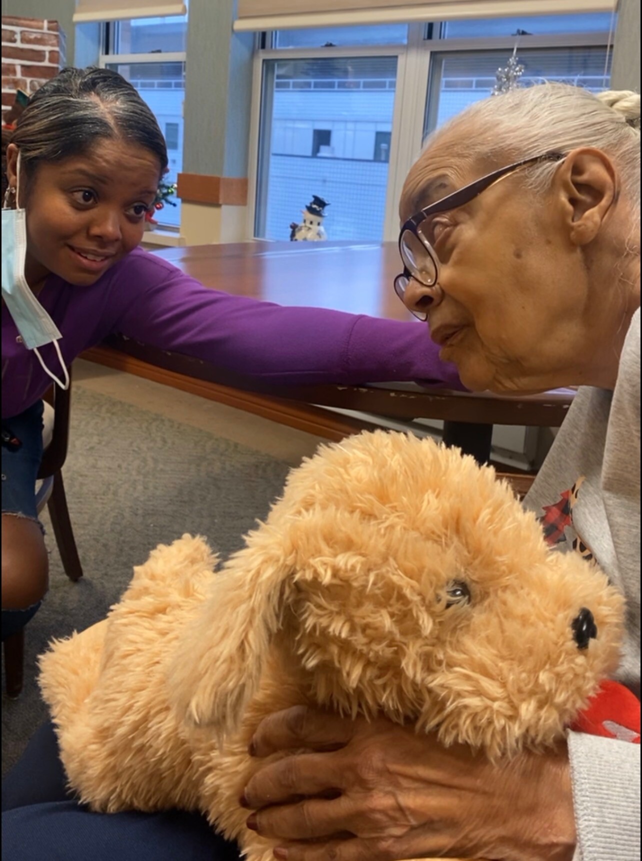 Young girl in a purple top leans over a table, speaking to an elderly woman with glasses; a fluffy tan teddy bear sits in the foreground.