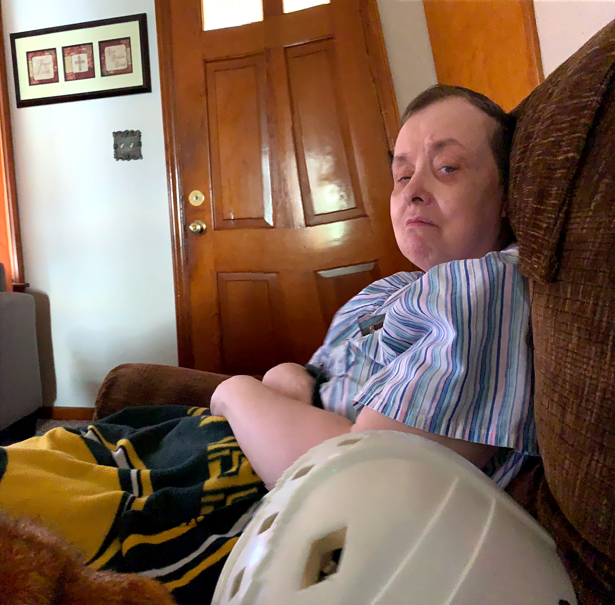 Person seated on a brown sofa in a living room, wearing a striped shirt, with a wooden door behind them and a patterned blanket nearby.