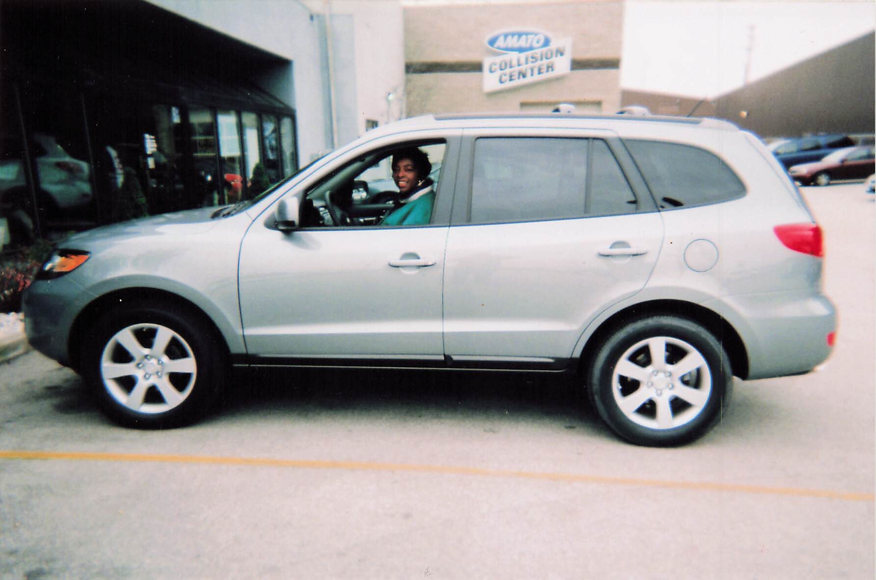 Smiling woman in a green top sits in the driver’s seat of a silver SUV parked in front of a building with a Collision Center sign in the background.