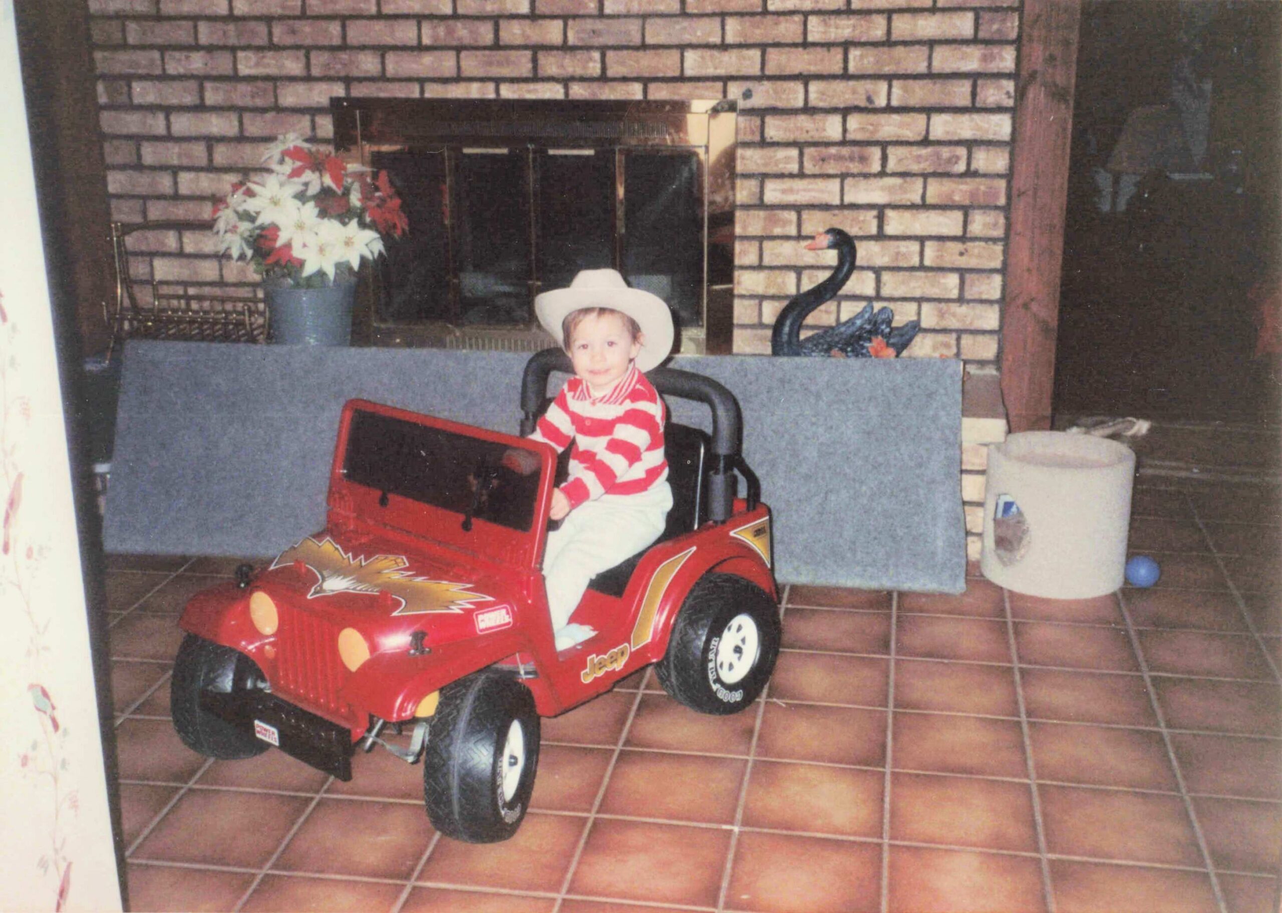 Child in a white cowboy hat rides a red toy Jeep in a living room with a brick fireplace backdrop.