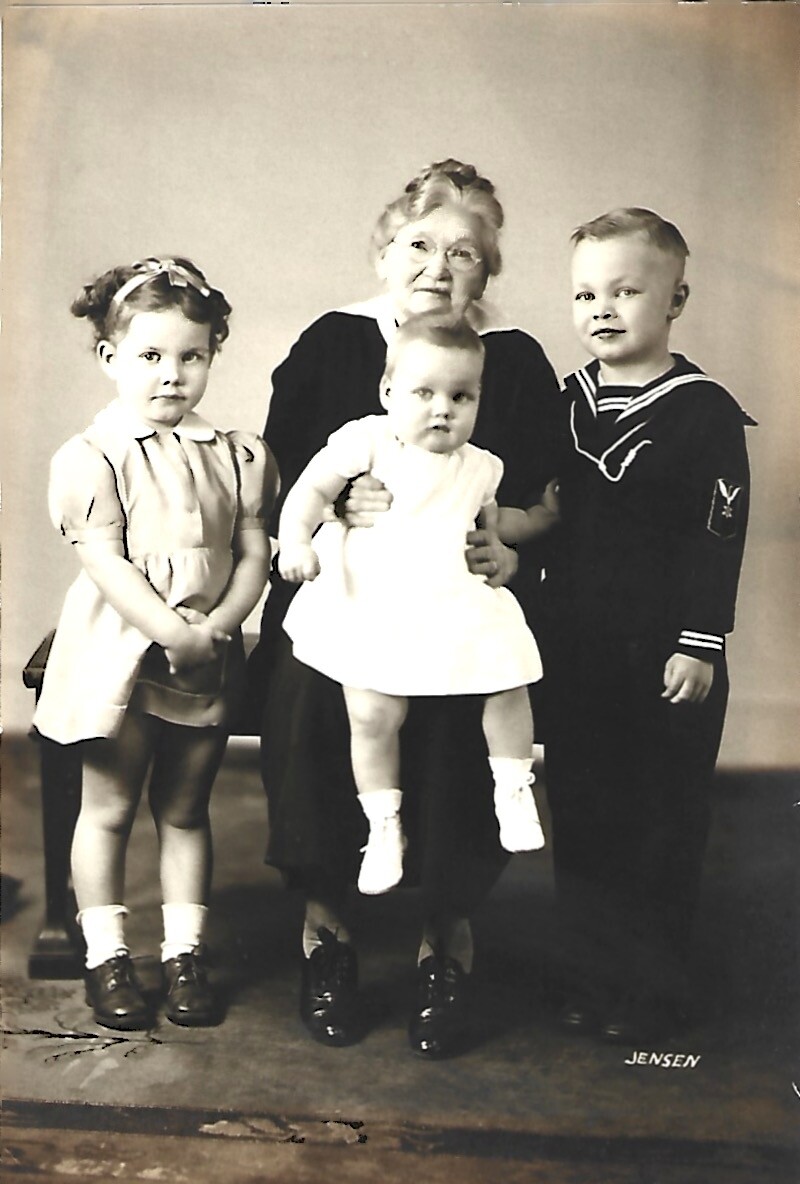 Elderly woman seated in the center holding a baby, with a girl on her left and a boy in a sailor suit on her right in a studio portrait.