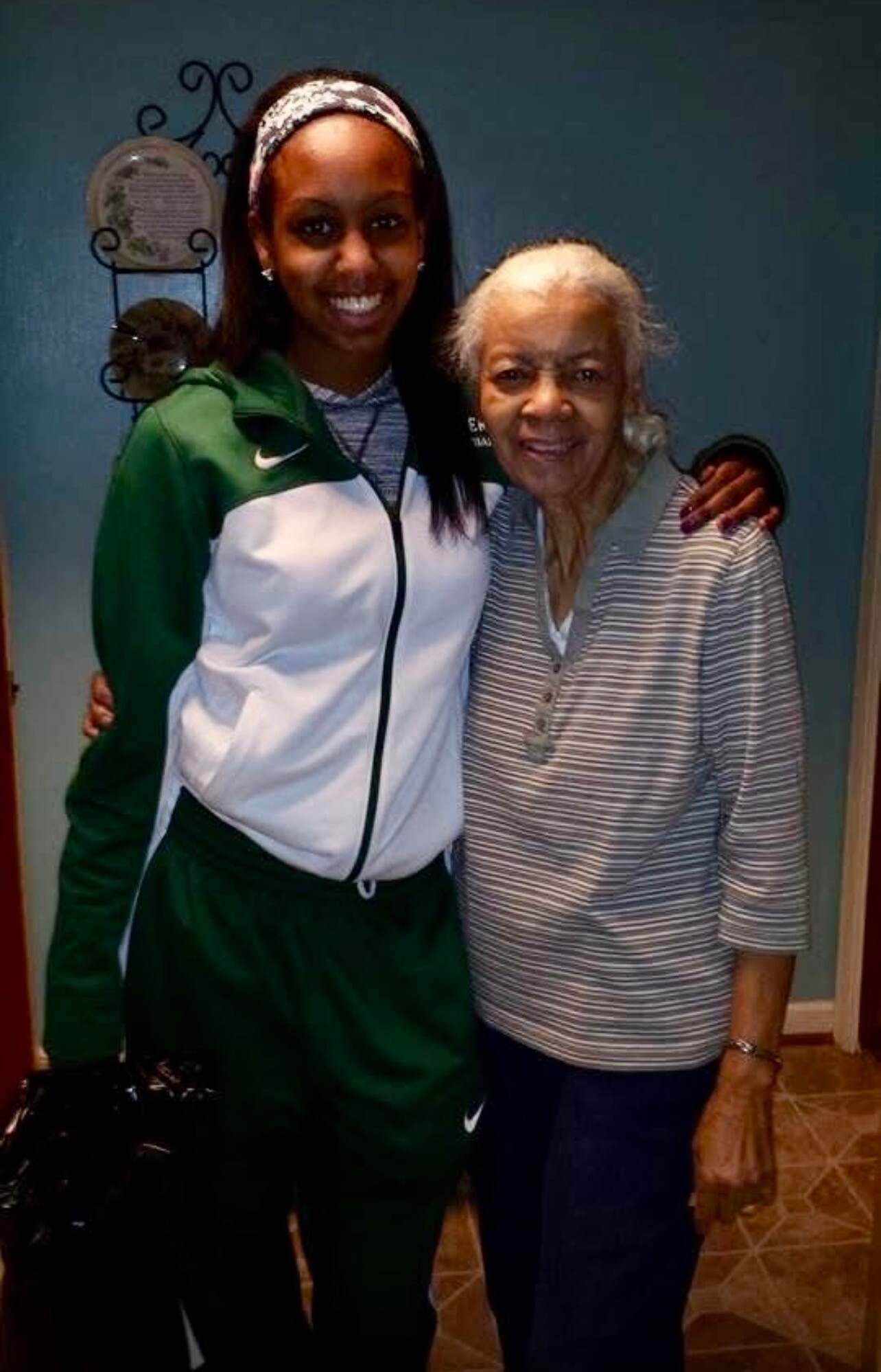 Two women posing together indoors, smiling warmly; younger woman in a green and white athletic jacket has her arm around an older woman in a striped shirt.