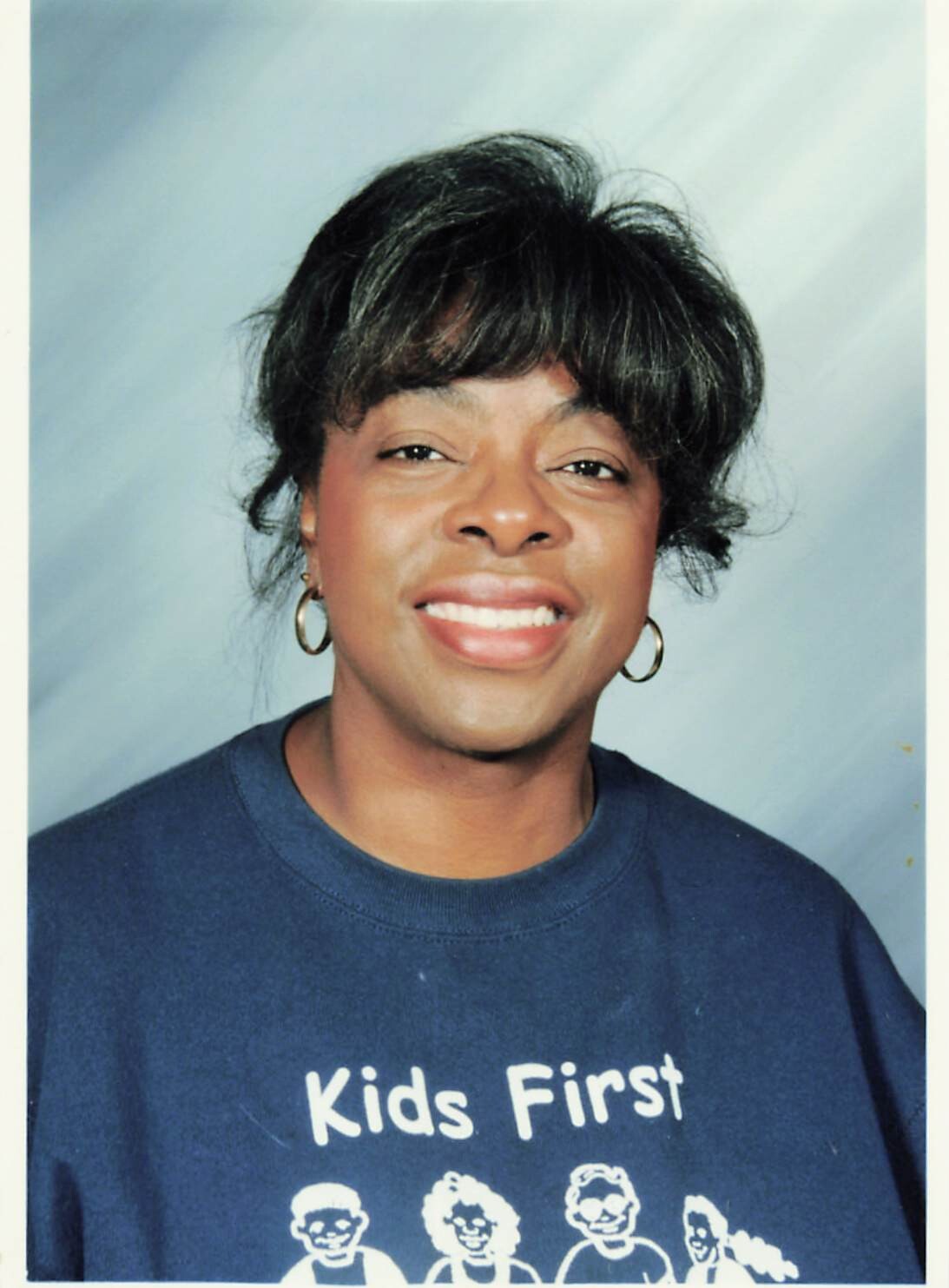 Smiling woman in a navy sweatshirt reading 'Kids First' with cartoon children graphic on the front.