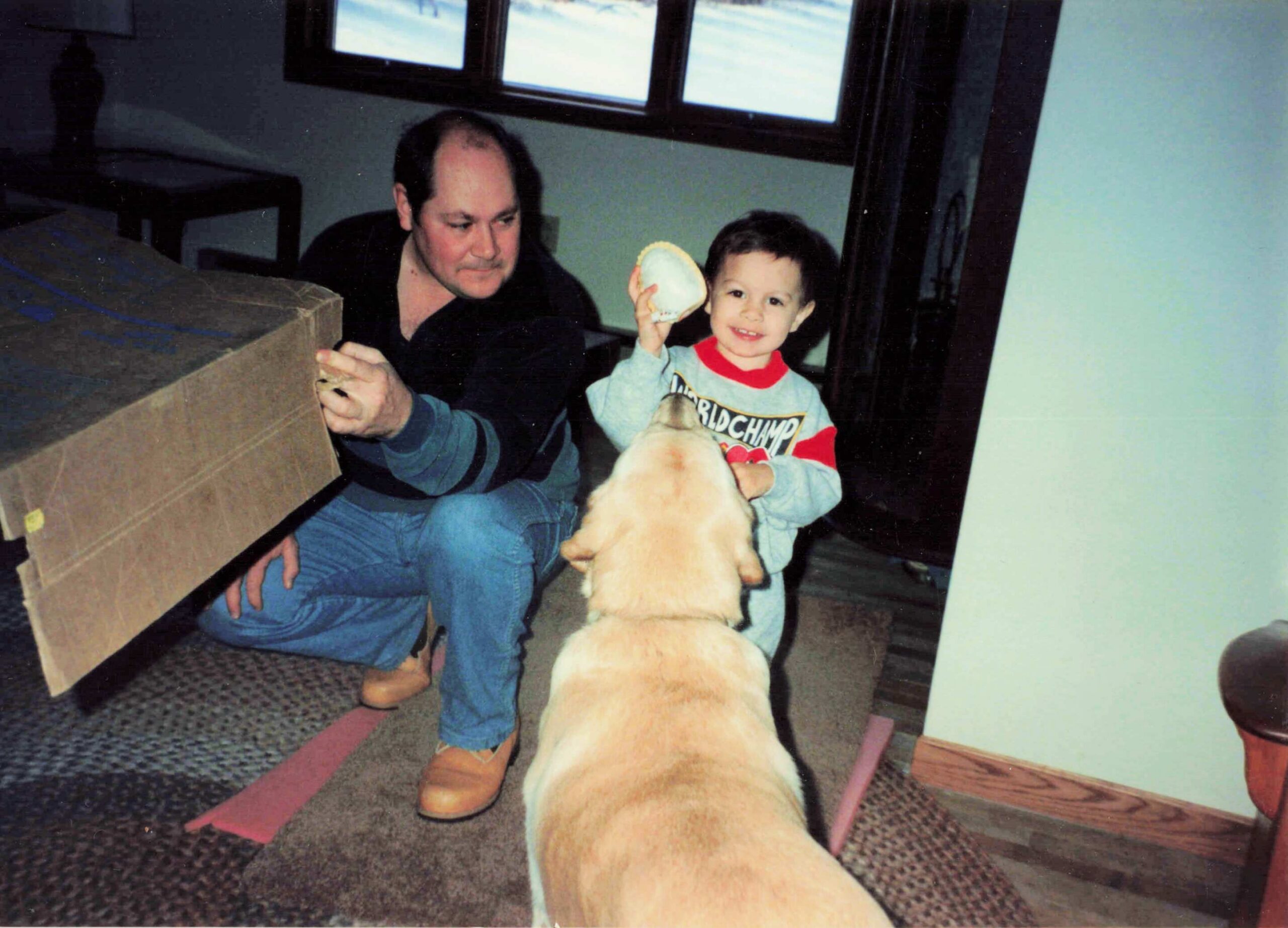 Father and young son kneel indoors beside a large cardboard box, with a beige dog in the foreground near the camera.