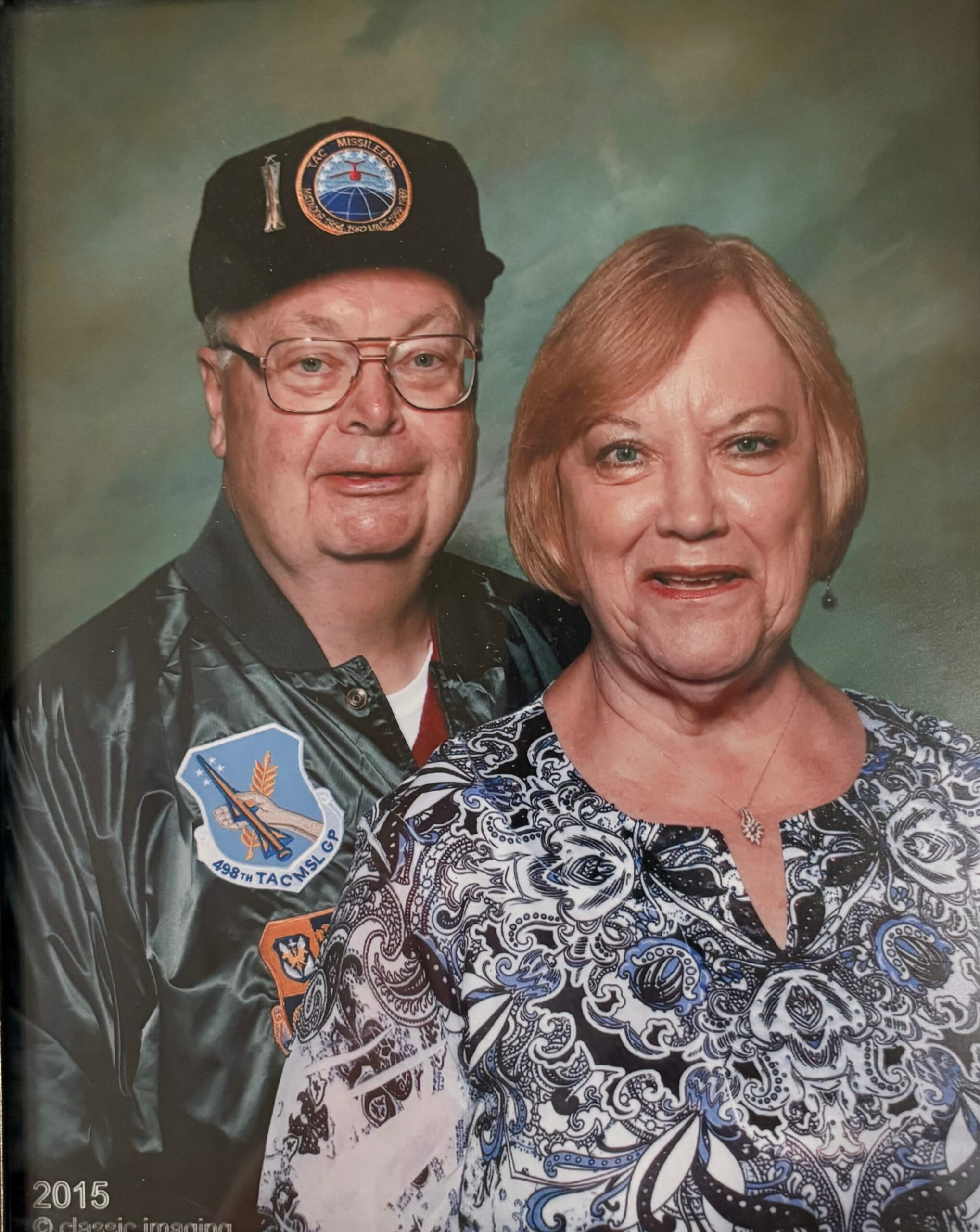 Portrait of an elderly man in a black cap with patches standing next to a smiling woman in a patterned blouse; '2015' visible in the corner.