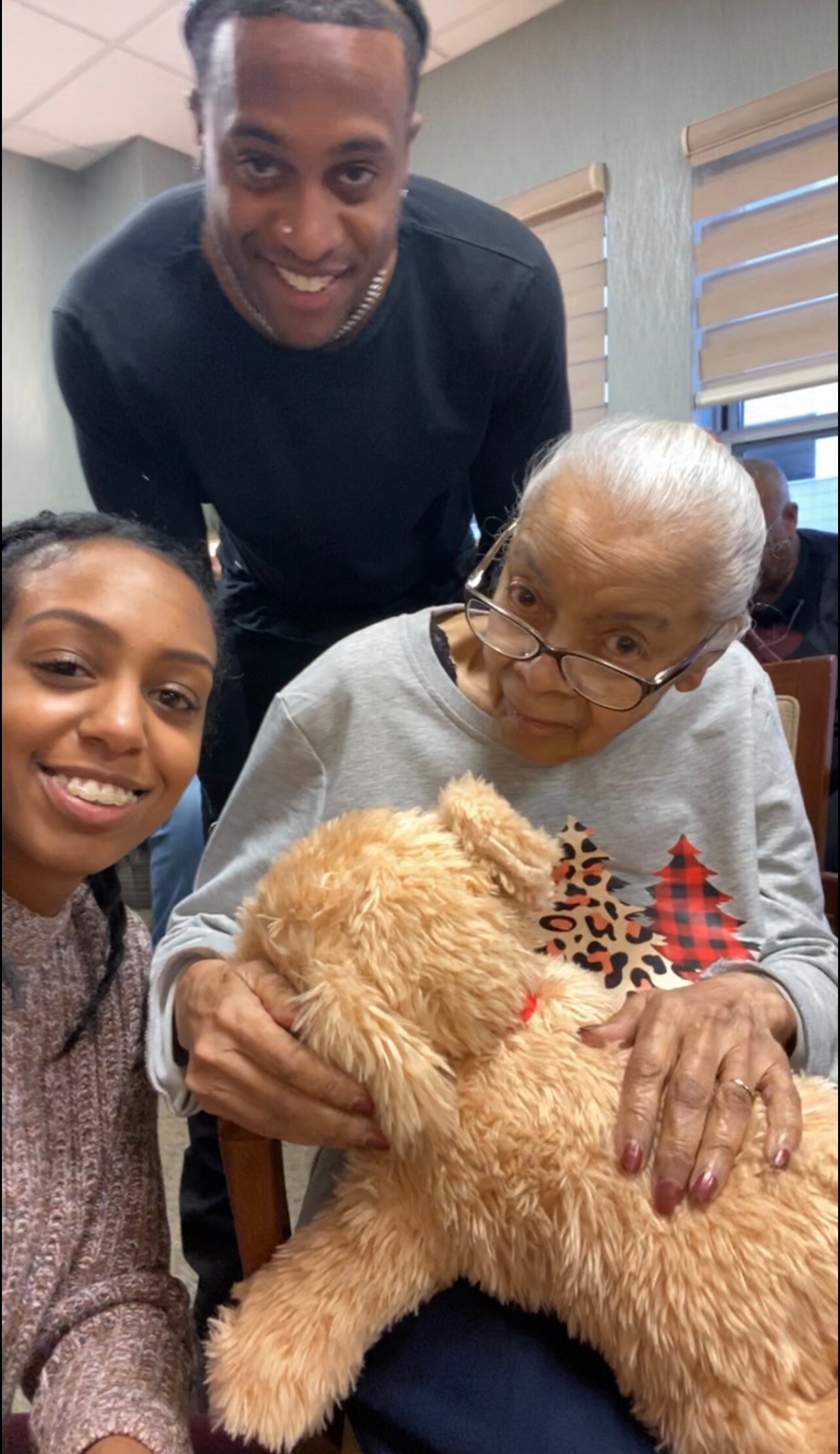 Young woman takes a selfie with an elderly woman holding a fluffy teddy bear while a smiling man leans in from behind.