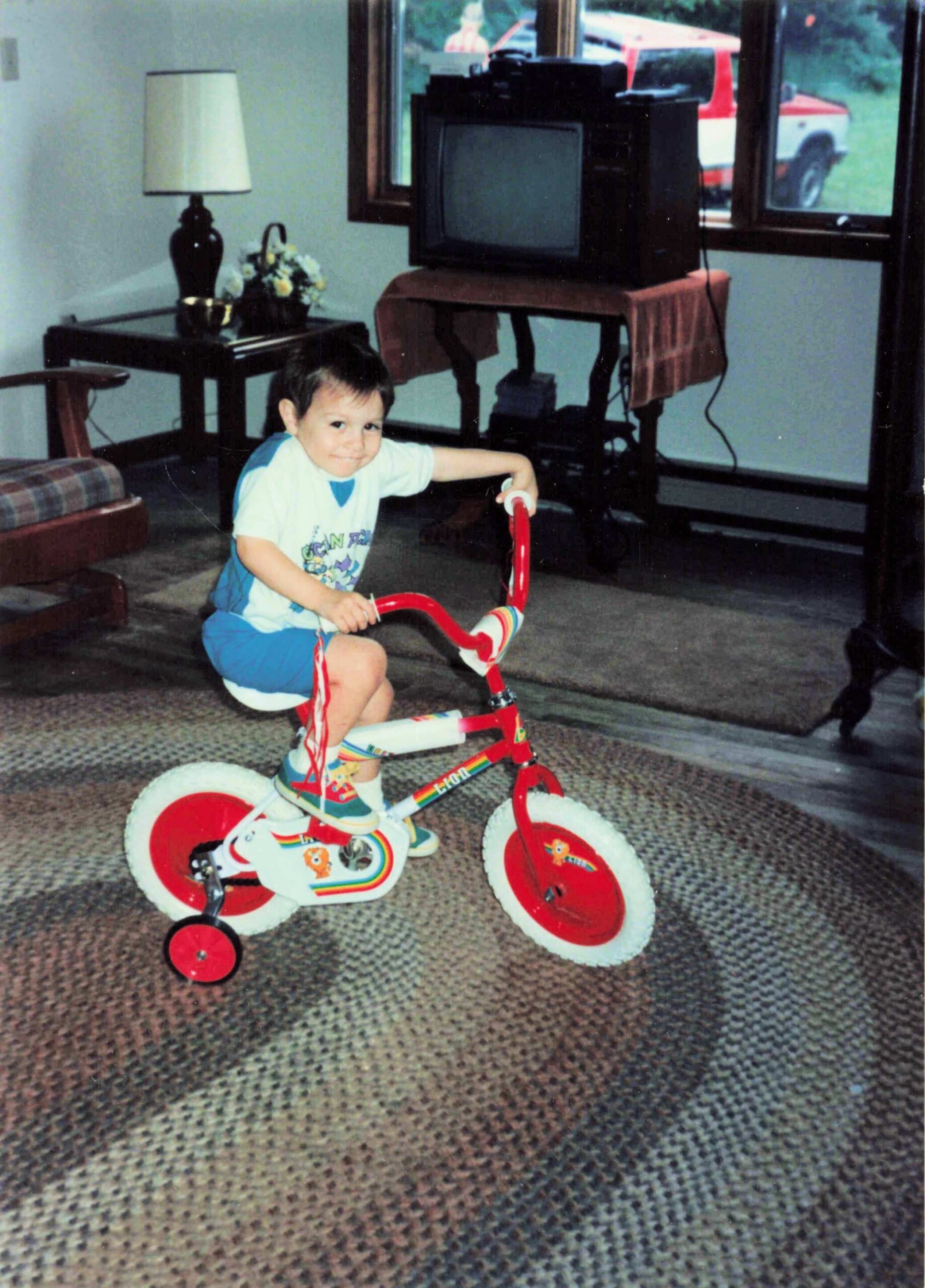 Smiling toddler riding a red tricycle with training wheels indoors in a living room, with a vintage TV on a table in the background.