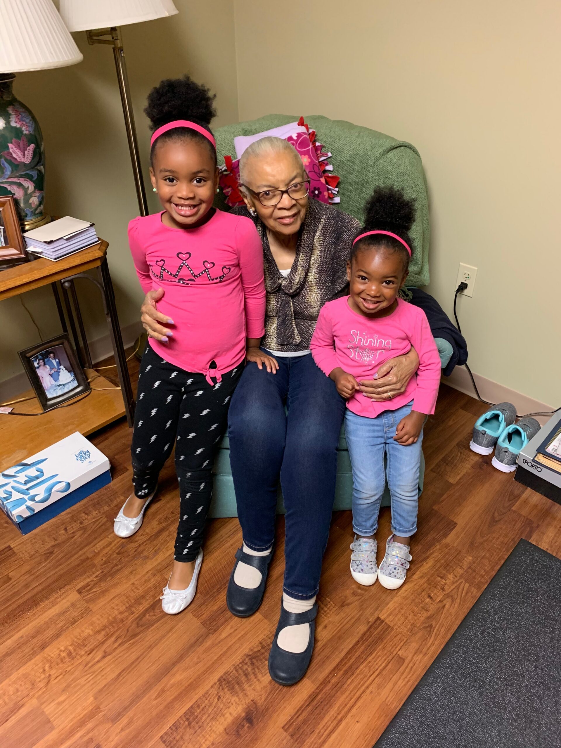 A smiling elderly woman sits in a green chair with two young girls in pink shirts on either side, indoors with a lamp and photo frames nearby (family moment).