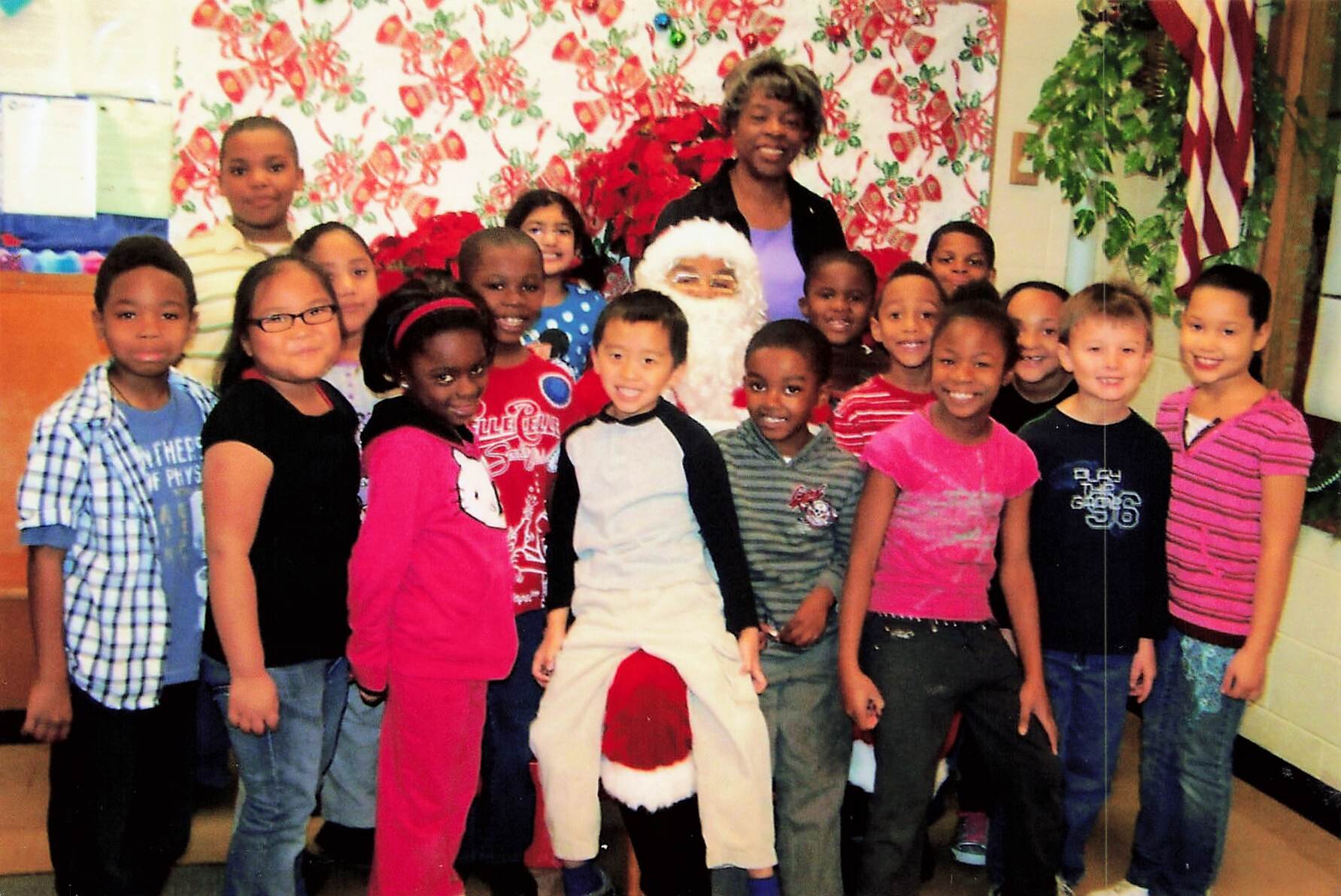 Group of smiling children posing with a person dressed as Santa Claus in a decorated classroom Christmas backdrop.