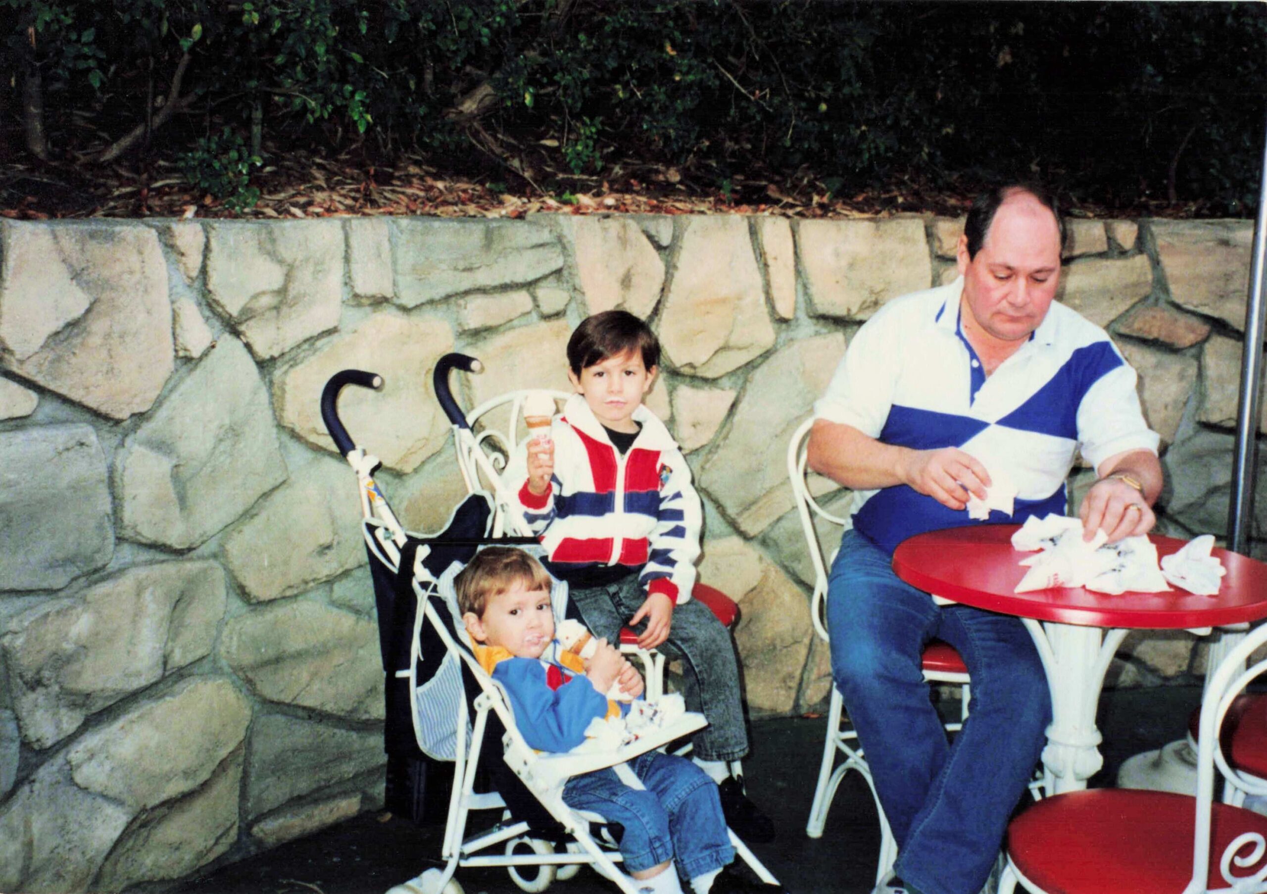 Father and two young children in strollers sit at red outdoor tables, eating ice cream beside a stone wall.