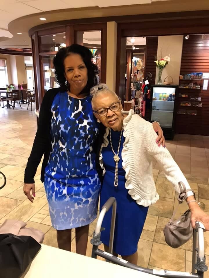 Two women stand together indoors, smiling for a photo: a younger woman in a blue animal-print dress and black cardigan beside an older woman in a blue dress with a white cardigan. The older woman uses a walker.