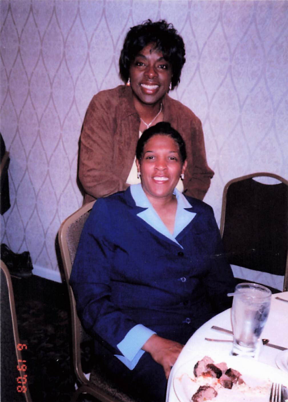 Two women smiling at a banquet table: one seated in a blue suit, another standing behind her in a brown jacket, with a plate of food and a glass nearby.