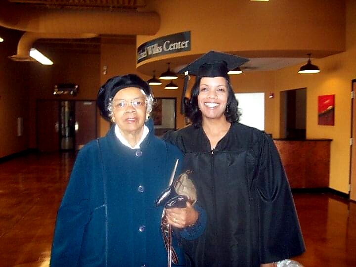 Graduate in cap and gown posing with an elderly woman in a blue coat in a campus hallway, both smiling.