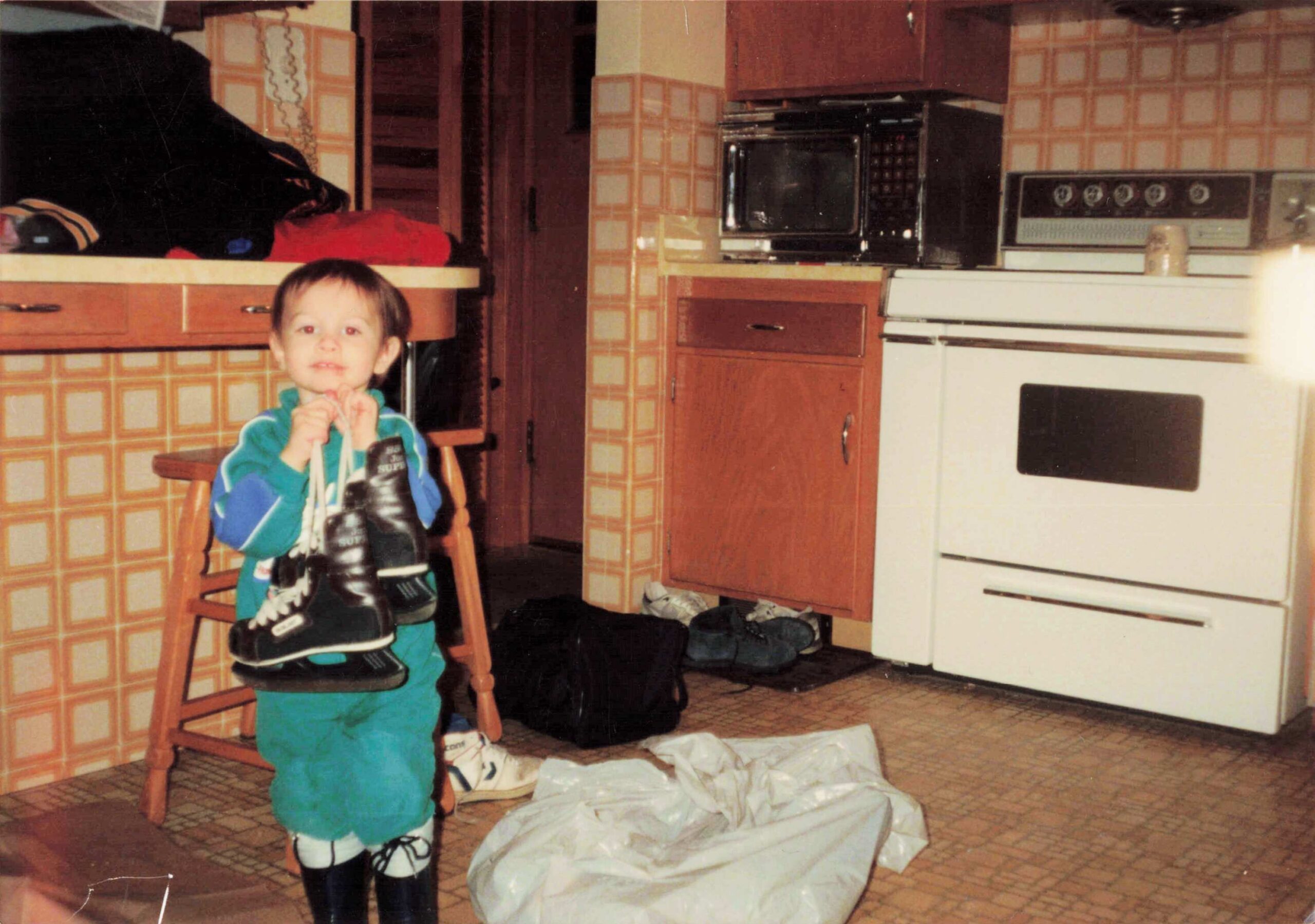 A young child in a turquoise outfit stands in a retro orange-tiled kitchen, holding a pair of black sneakers with white laces, while clothes and bags lay on the chair and floor nearby.