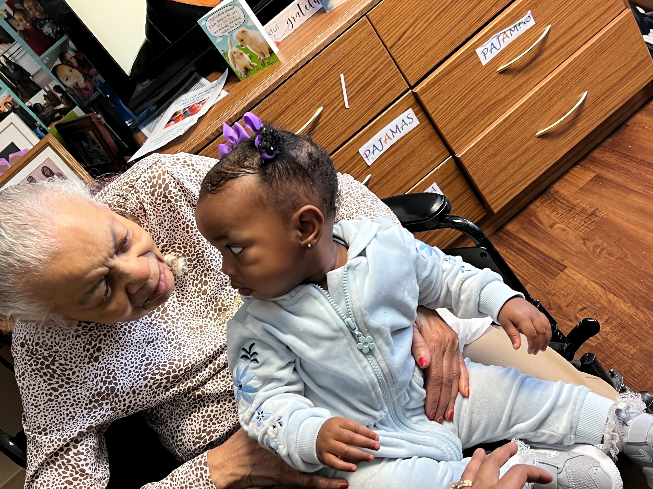 Older woman and baby sharing a tender moment, seated together in a room with wooden drawers in the background.