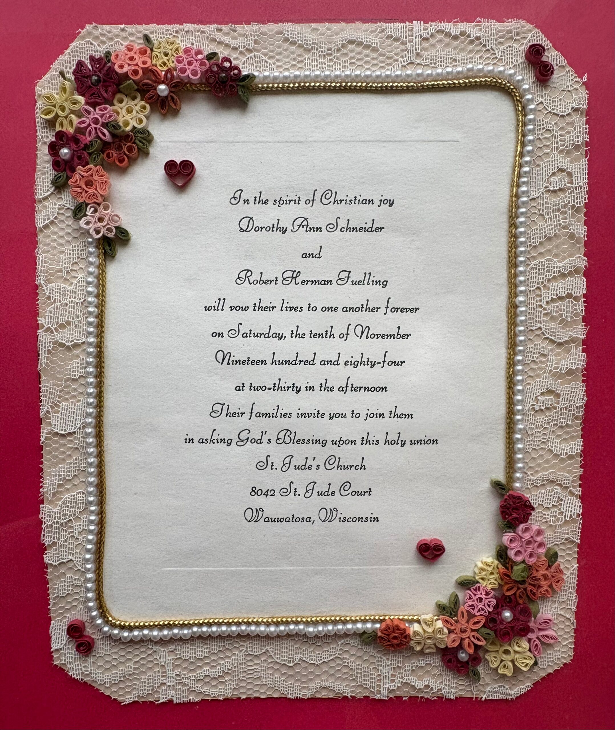 Decorative wedding invitation framed by lace, pearls, and rolled paper flowers on a red background, with scripted text panel in the center.