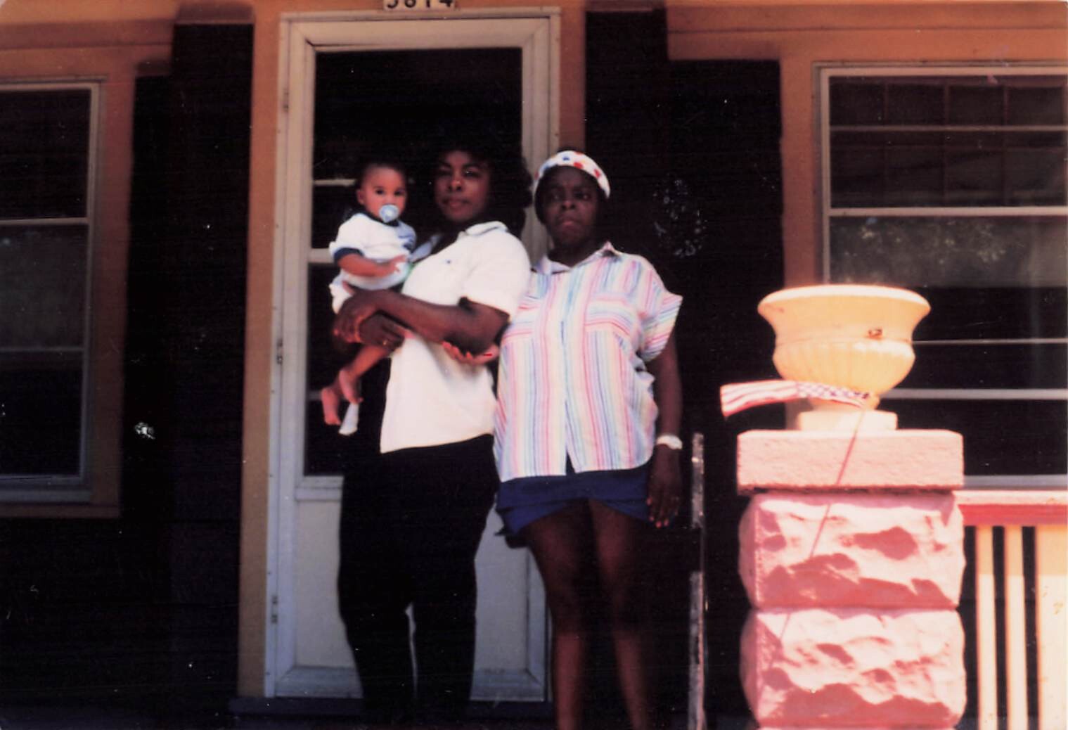 Two women stand on a front porch with a baby; one woman holds the infant while the other stands beside them by the doorway of a house with a decorative planter nearby.