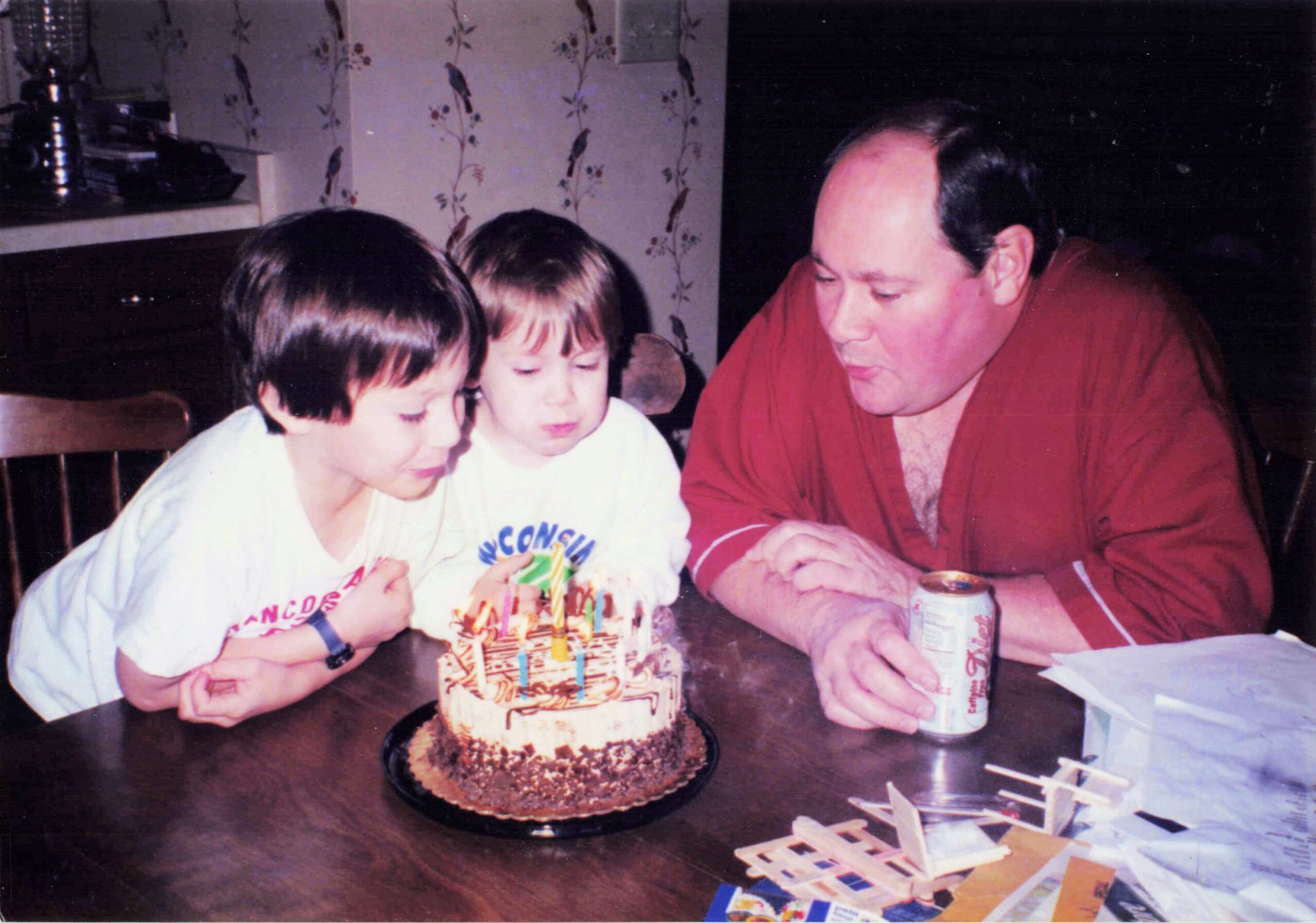 Family at a kitchen table celebrating a birthday, with two kids and a man in a red shirt around a lit cake covered in colorful candles and decorations on the table.