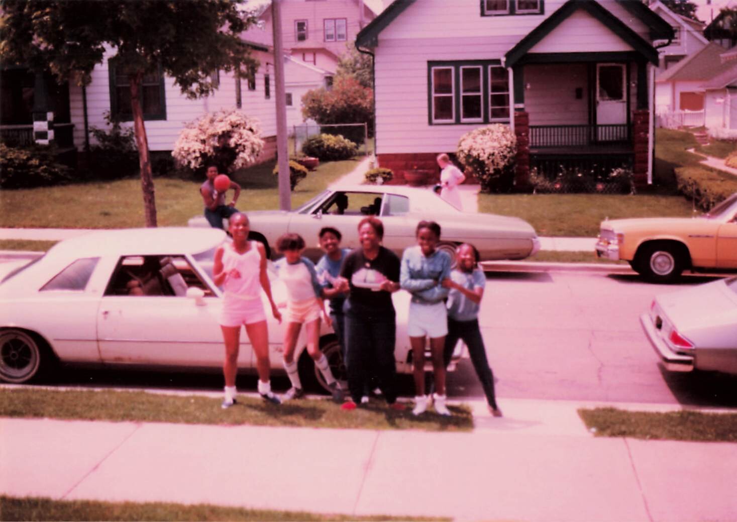 Group of friends posing together in a suburban street, standing in front of several parked cars with houses and lawns in the background