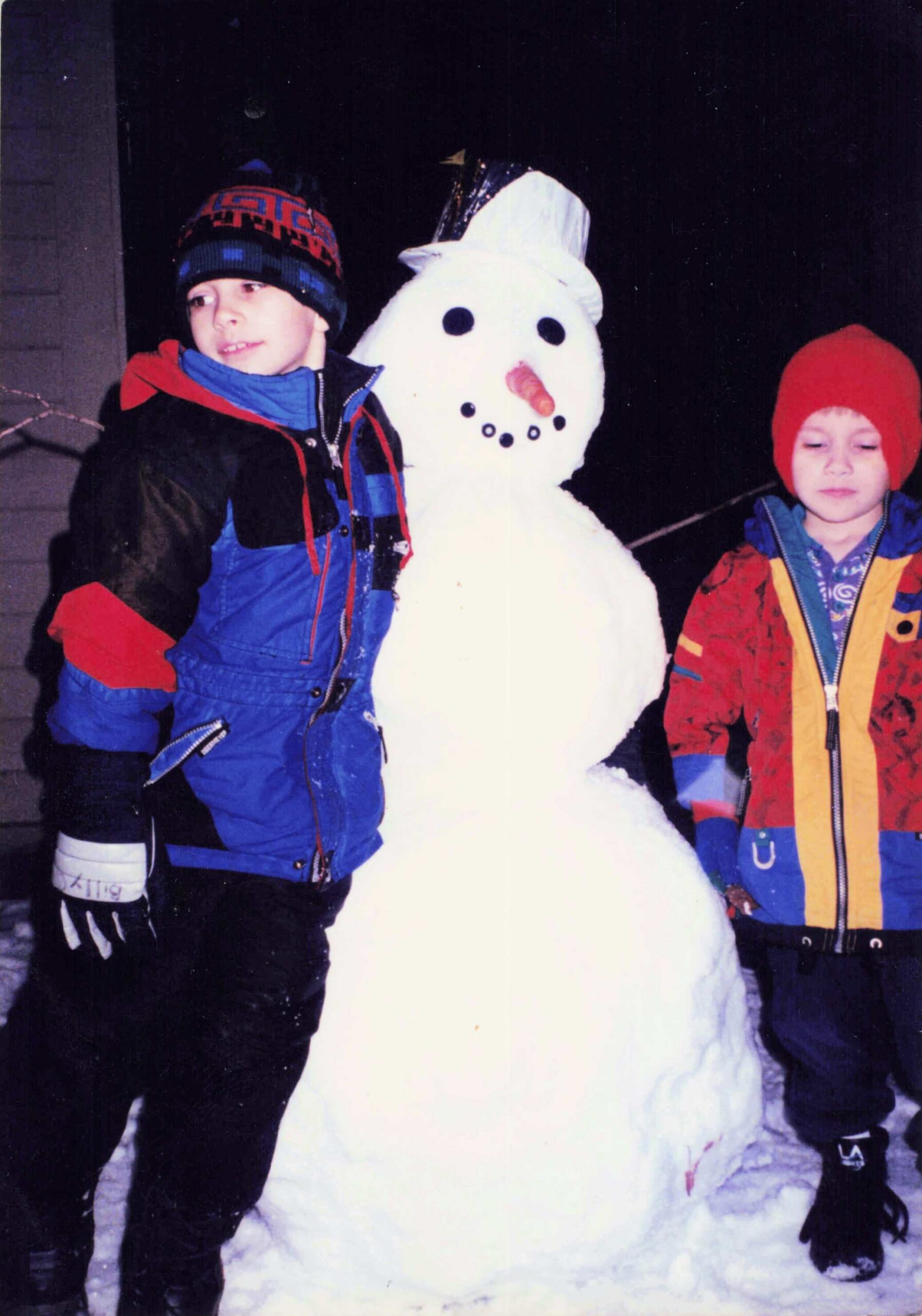 Two kids in colorful winter jackets pose with a tall snowman wearing a top hat and carrot nose at night in a snowy scene.