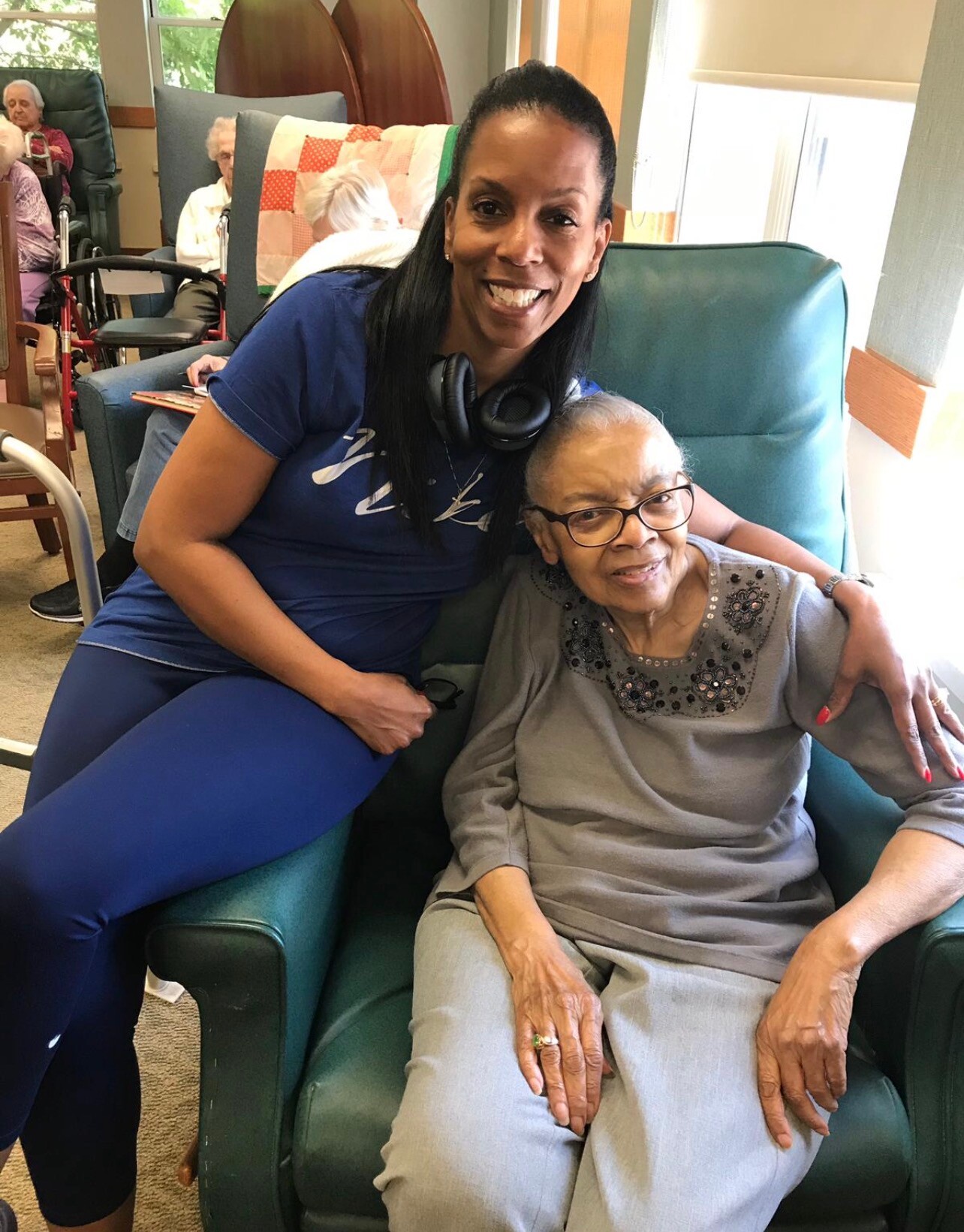 Smiling caregiver in blue scrubs sits beside an elderly woman in a teal chair, both smiling in a care home lounge.