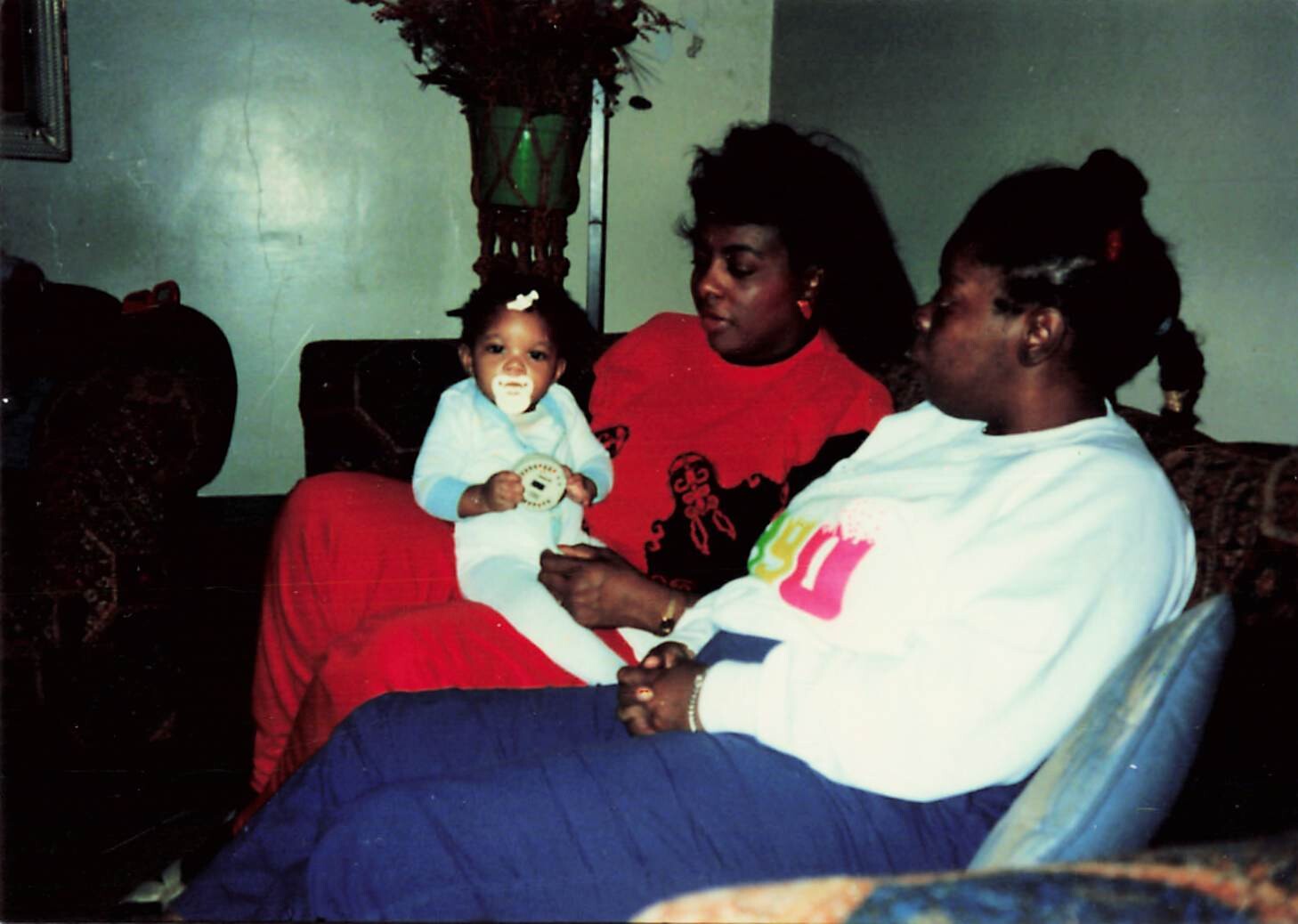 A baby with a pacifier sits on a woman in a red shirt, while another woman in a white sweatshirt sits nearby in a living room.