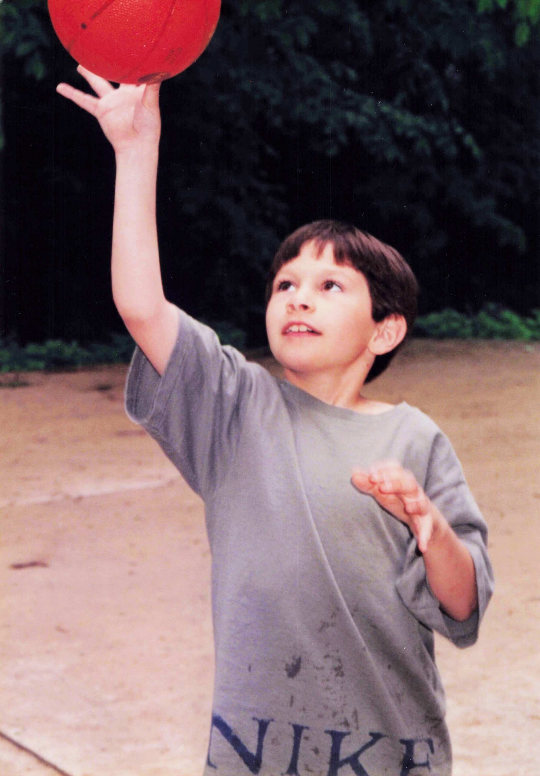 Child reaching up to catch a red ball outdoors, on a dirt field with trees in the background