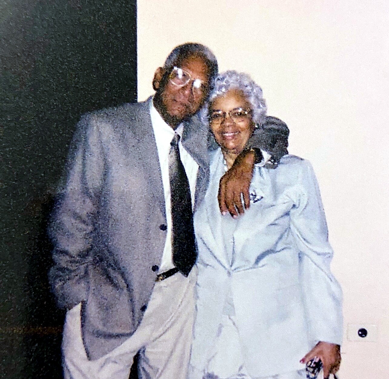 Smiling older man in a gray suit with his arm around a smiling older woman in a light gray blazer, posing together indoors.
