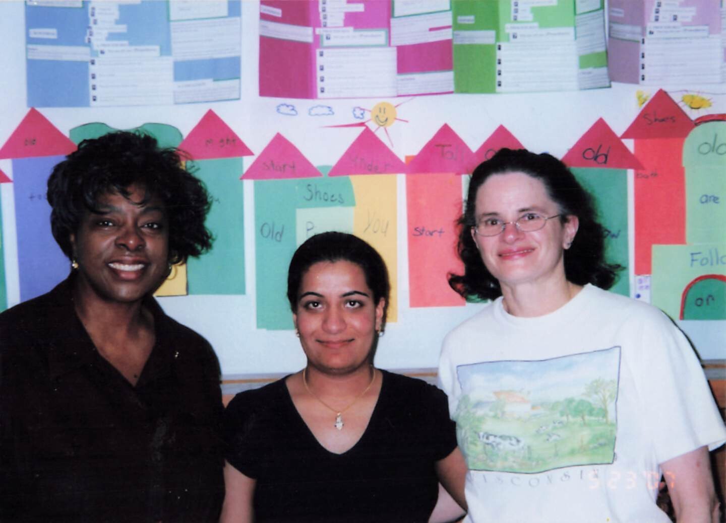 Three women posing together in a classroom with colorful wall charts and paper shapes behind them.