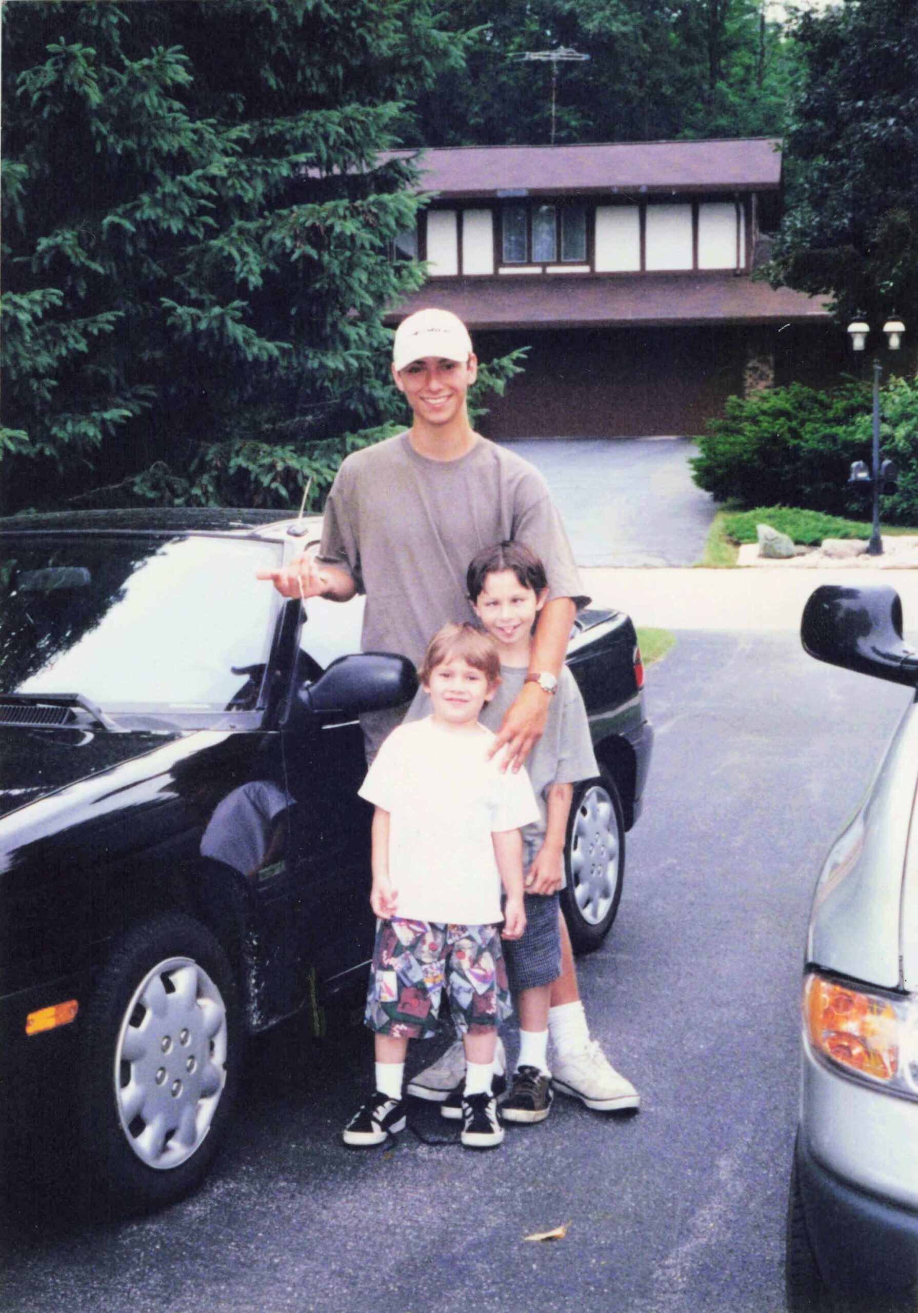 Smiling man in a baseball cap stands with two children by a dark car on a residential driveway, house and trees in back.