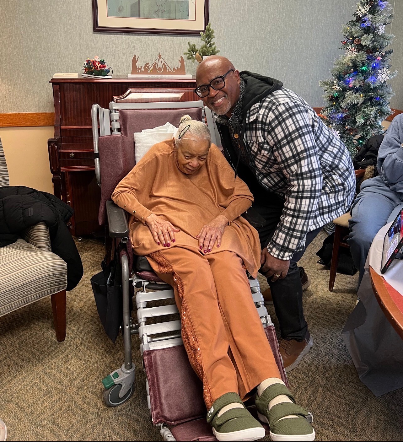 Smiling man leans beside an elderly woman in a wheelchair in a holiday-decorated room.