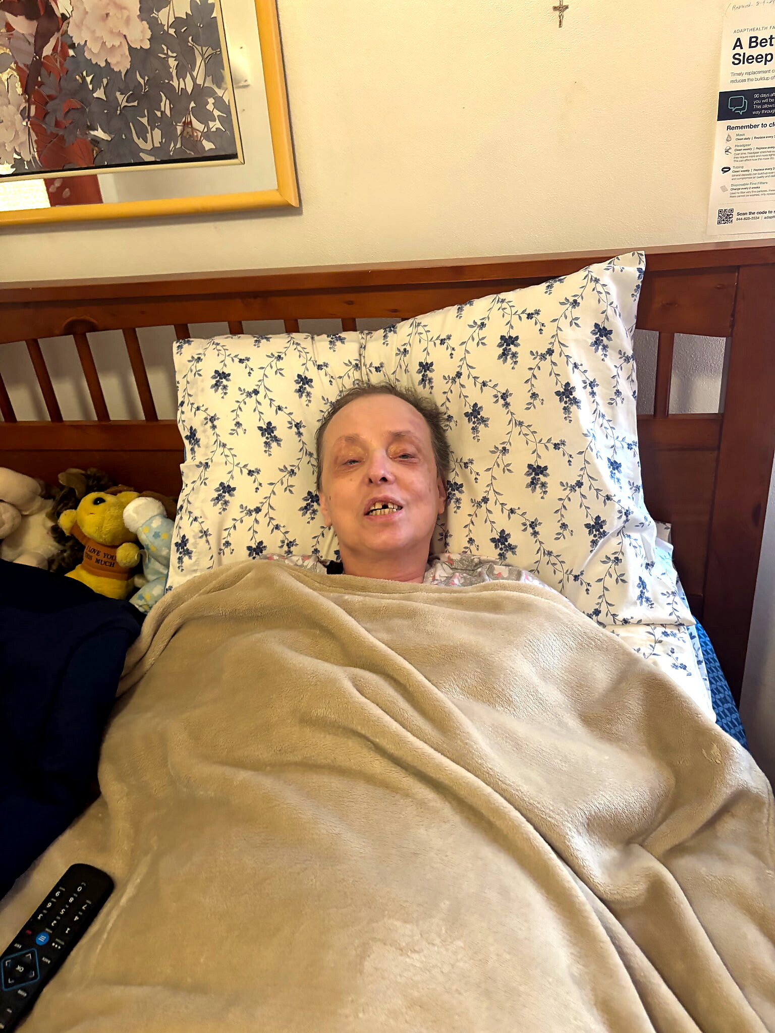 Person lying in bed, head on a floral pillow, covered with a beige blanket, with a remote nearby and stuffed toys visible at the side.