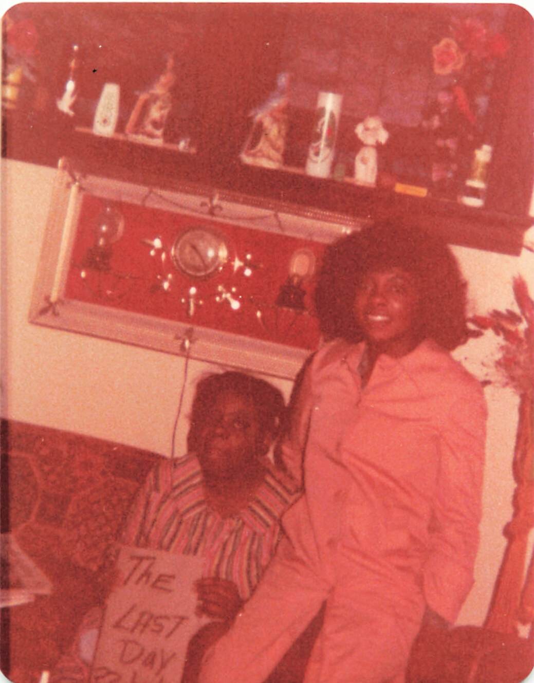 Two women pose in a living room: a woman in pink stands beside a seated woman in a striped shirt, holding a poster reading 'THE LAST DAY'.