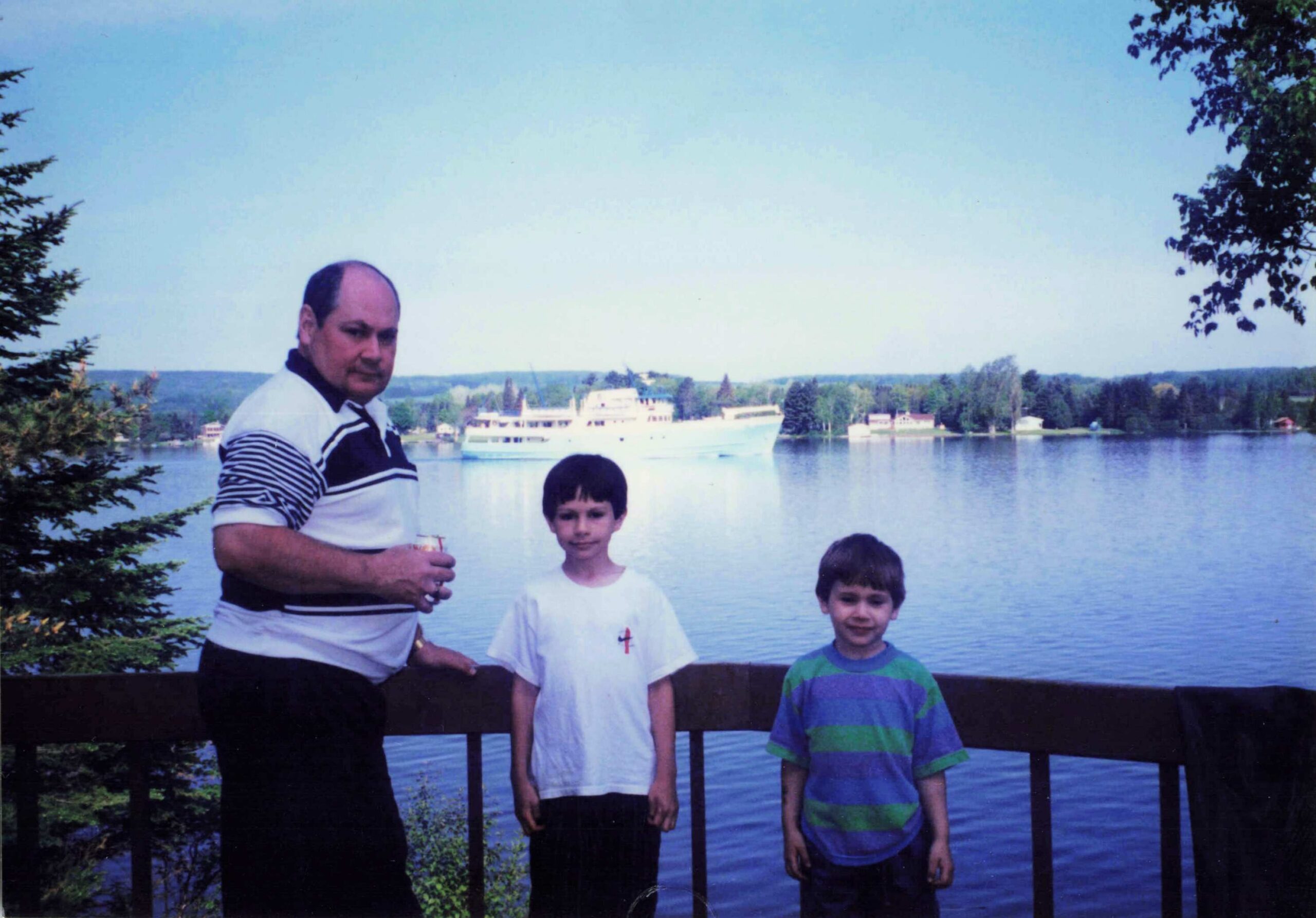 Man and three children posing by a wooden railing along a lake, with a large white boat in the background on a sunny day.