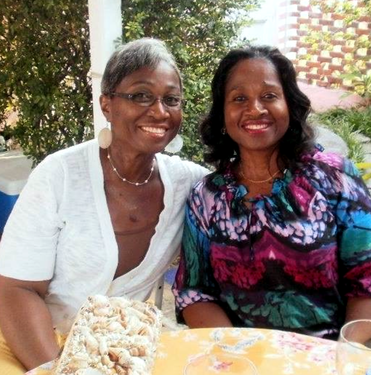 Two women smiling at an outdoor table, with a bag of seashells on the table in front of them.