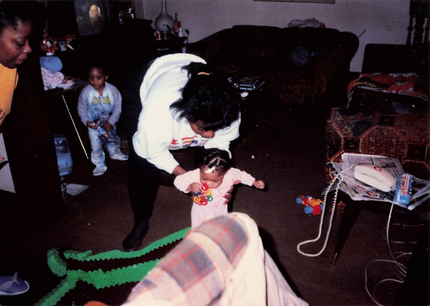 Caregiver helping a toddler walk across a cluttered living room while a young child looks on.