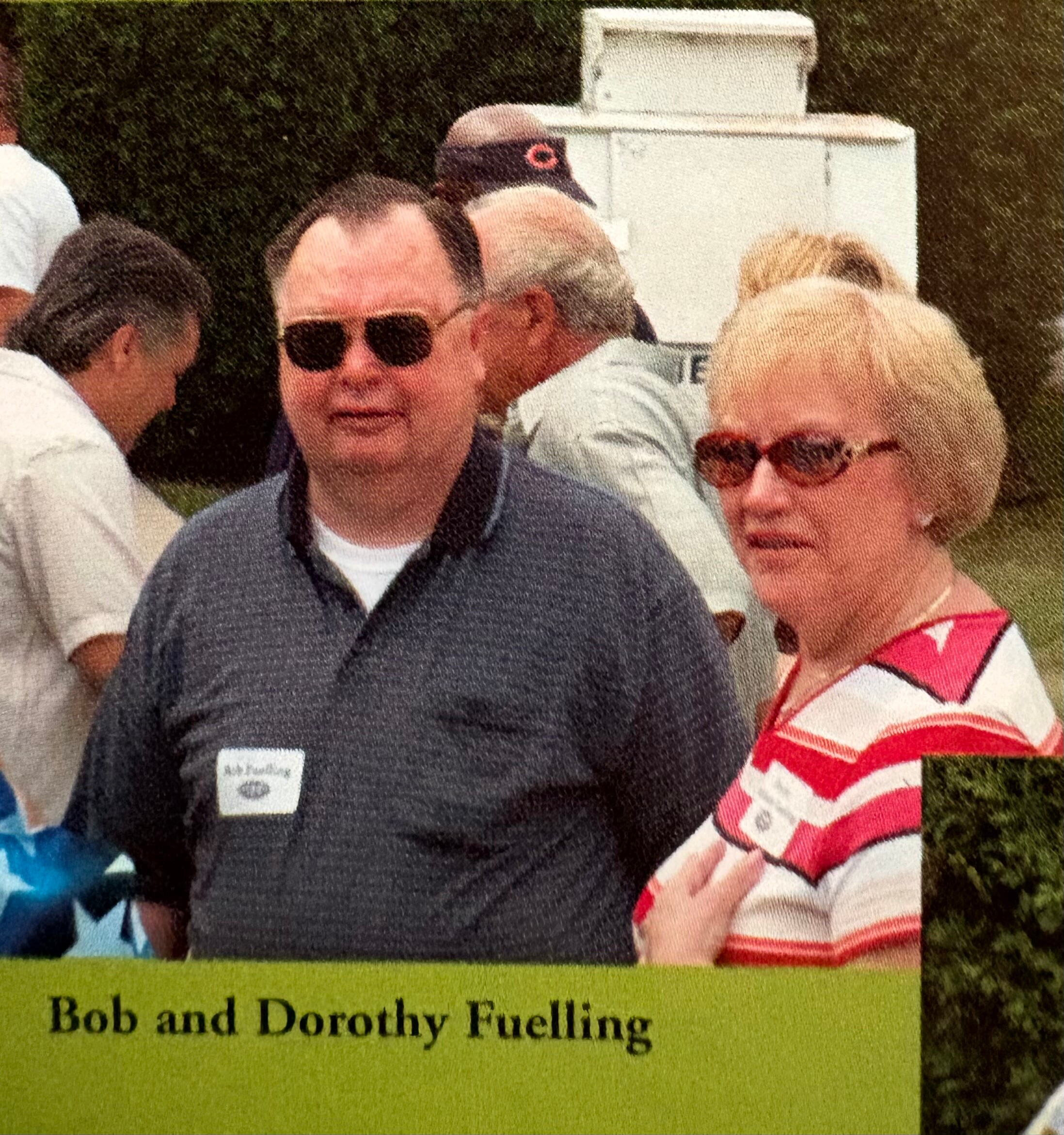 Group of people posing outdoors at an event; man in a dark polo and sunglasses in the foreground, woman in pink striped top beside him; green caption reads 'Bob and Dorothy Fuelling'