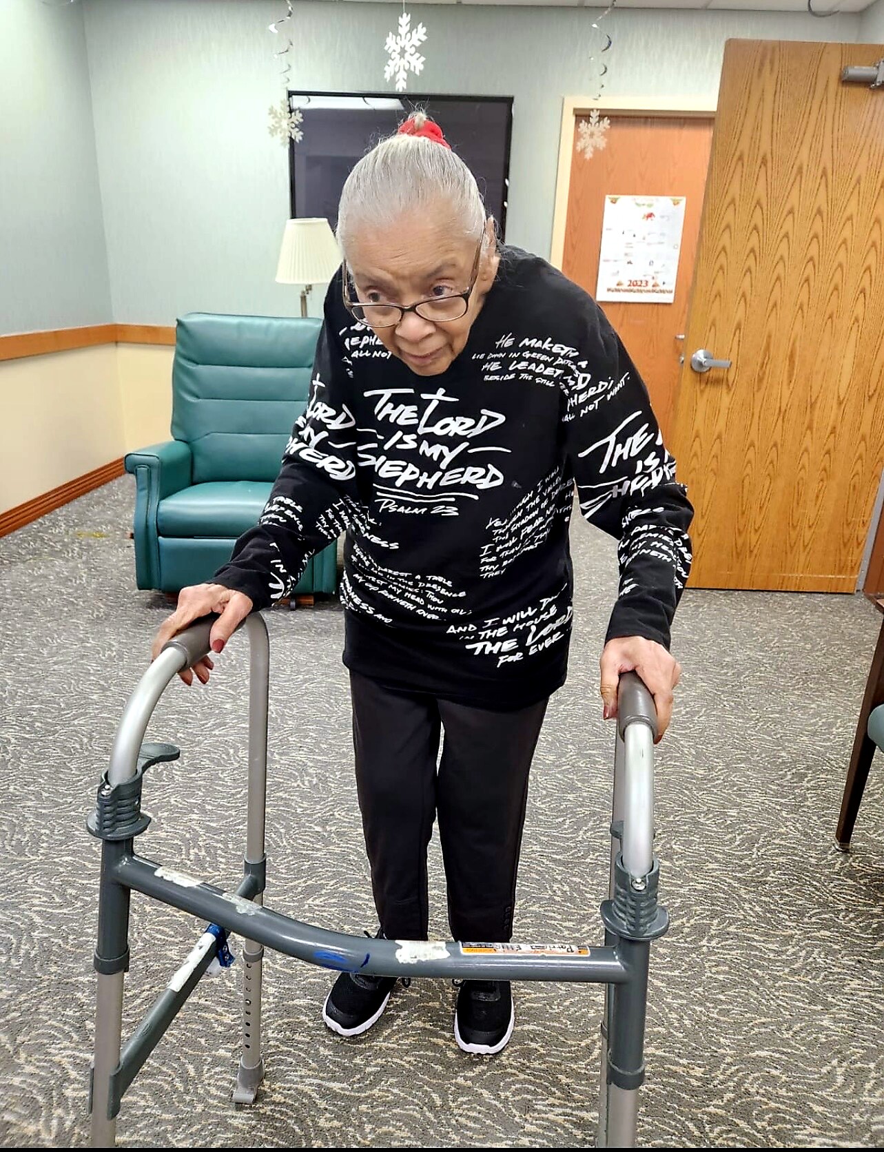 Elderly woman with gray hair in a bun using a walker in a room with hanging snowflake decorations. Note: The second line is another option if needed.