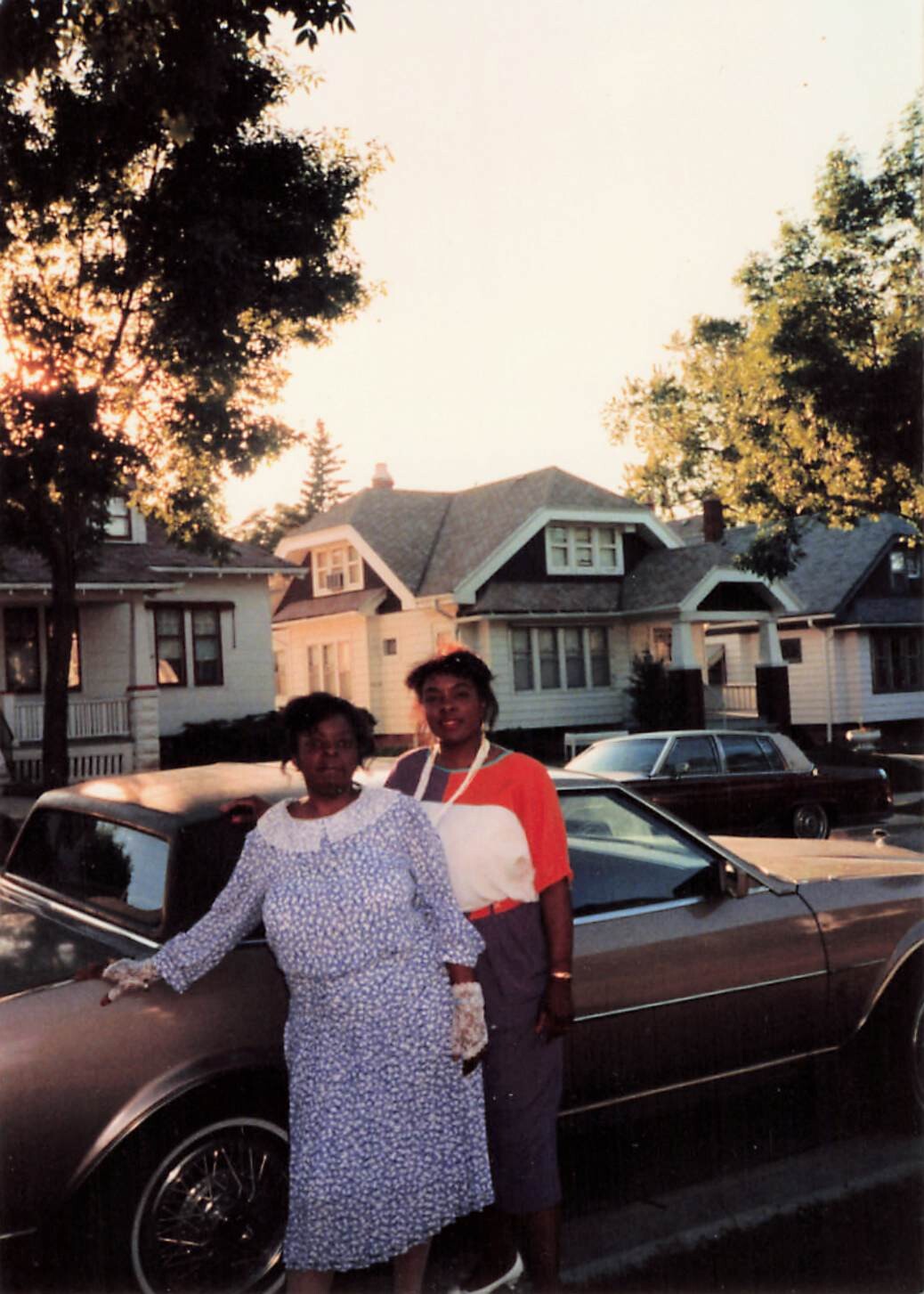 Two women posing beside a vintage car on a suburban street at sunset with 1950s–60s houses in the background