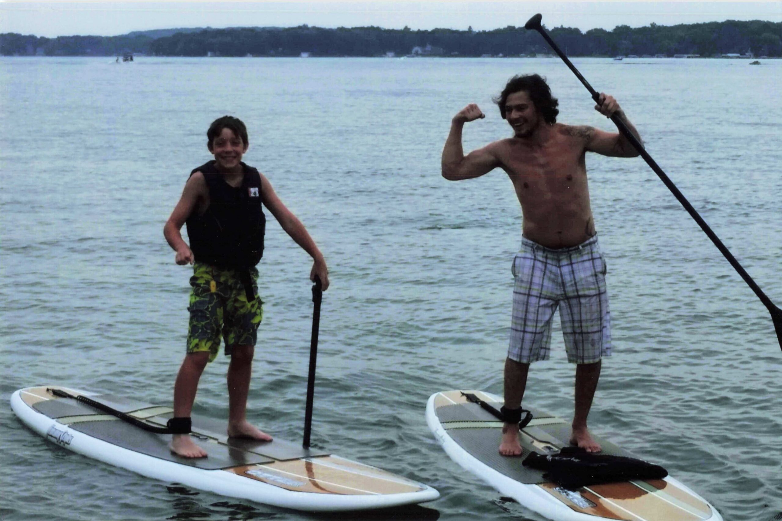 Two people stand on stand-up paddleboards in shallow water near shore, smiling at the camera; one boy wearing a black life vest, the other shirtless with plaid shorts and a long paddle raised.