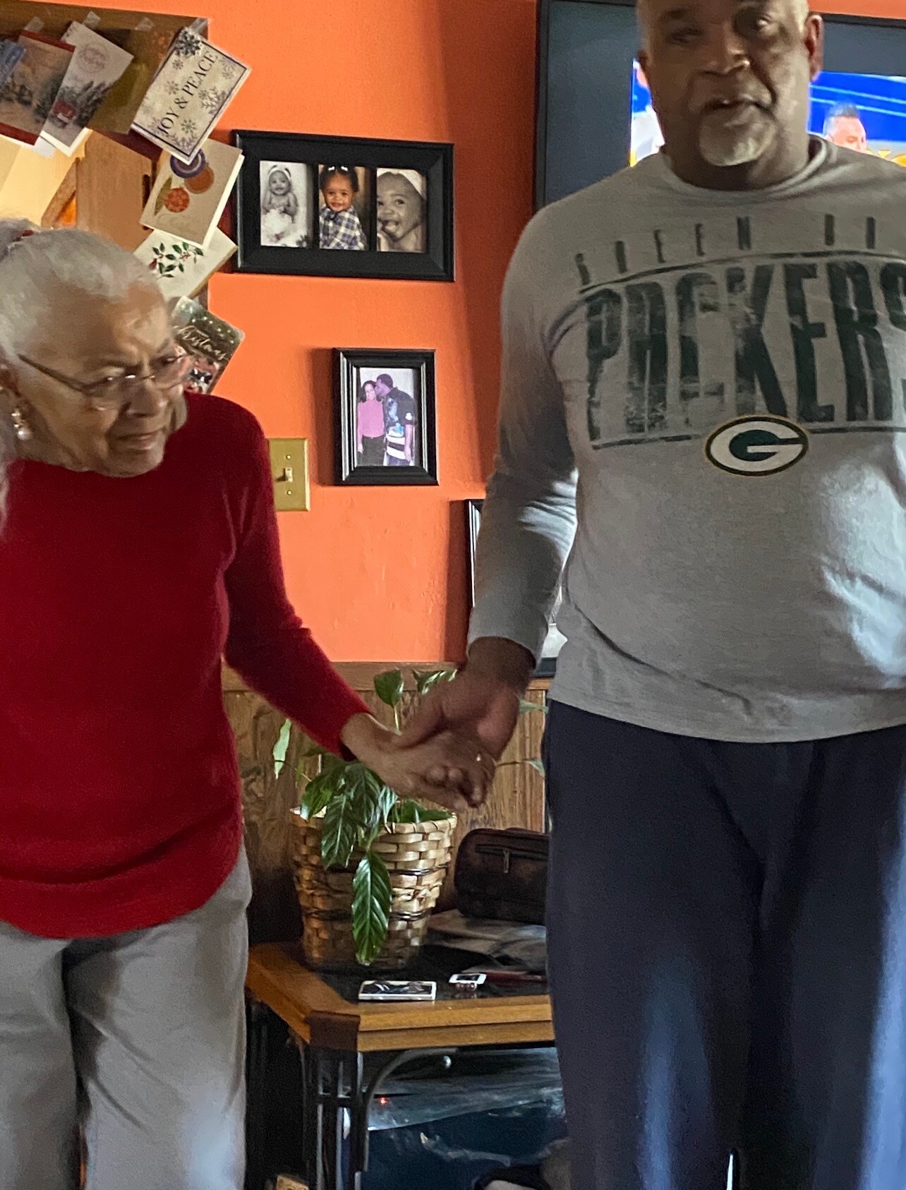 An elderly woman in a red sweater and a man in a gray Packers shirt hold hands in a living room with orange walls, family photos, and a plant in the background.
