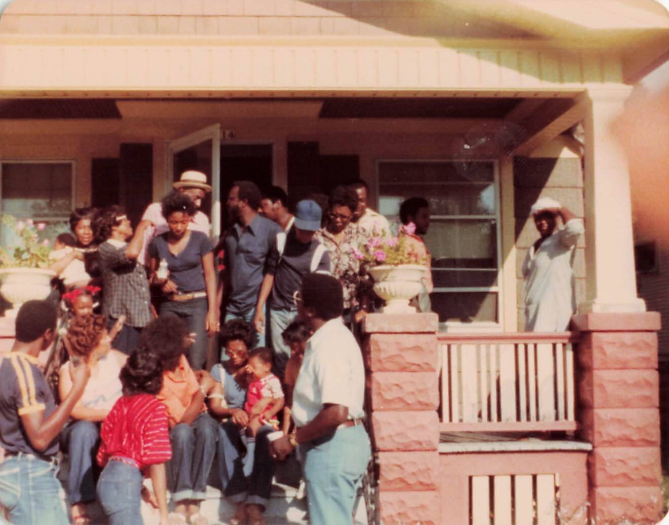 Large family gathering on the front steps of a yellow house, with adults and children chatting and posing for a photo.