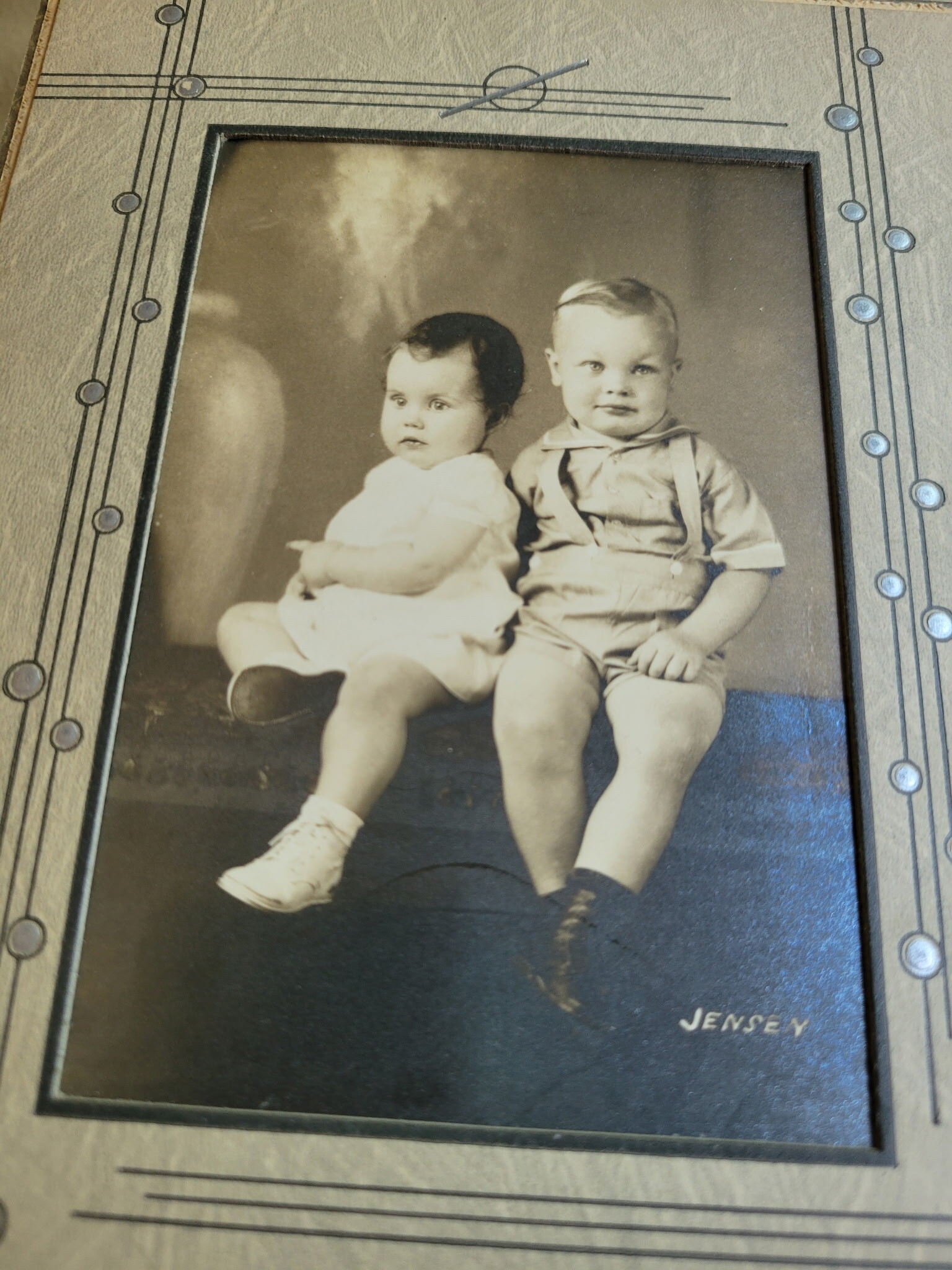 Two young children sit together for a studio portrait; girl in white dress on left, boy in light outfit with suspenders on right, framed photo with a Jensen signature.