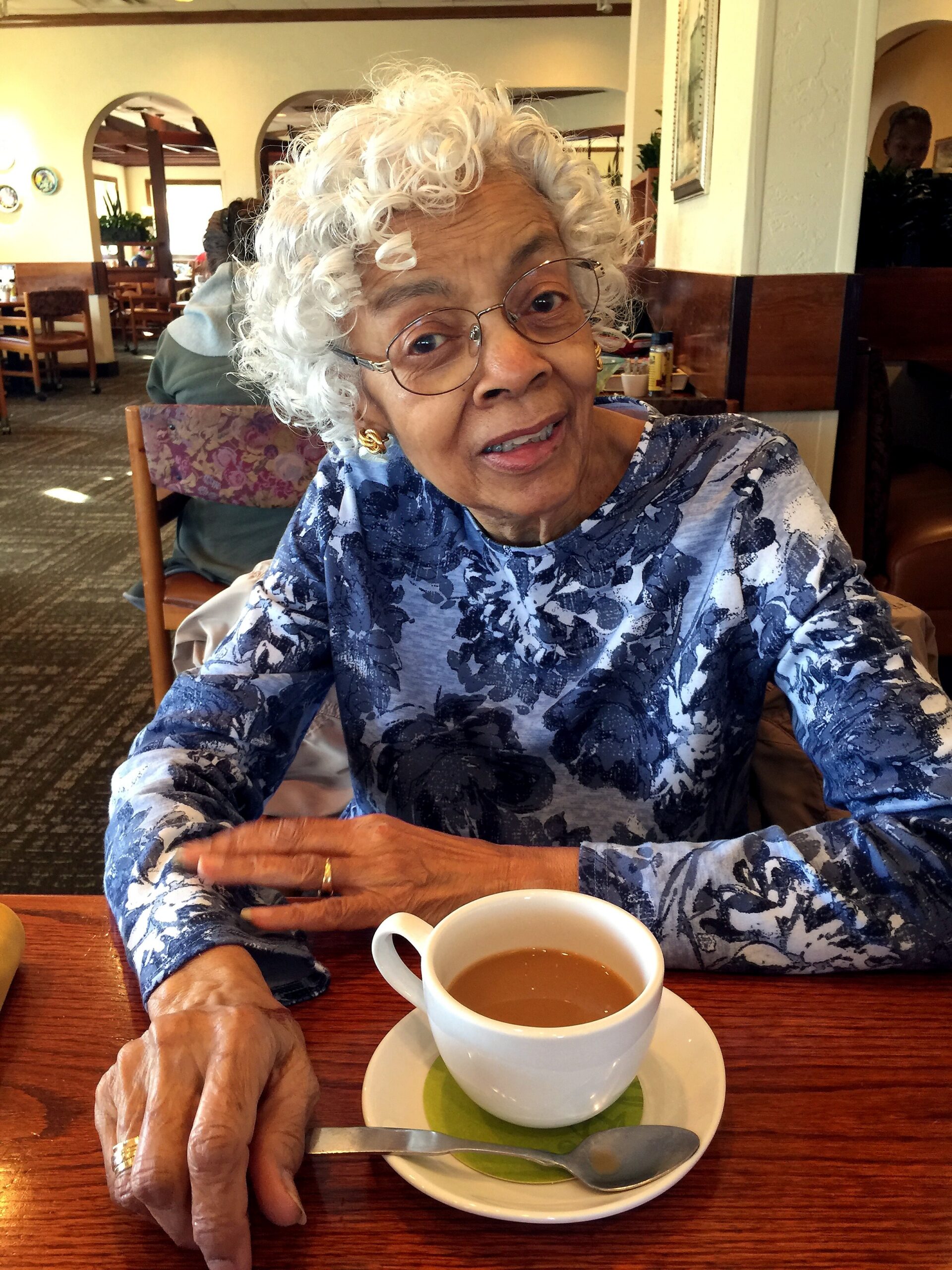 Elderly woman with white curly hair and glasses sits at a restaurant table, a cup of tea on a saucer in front of her.
