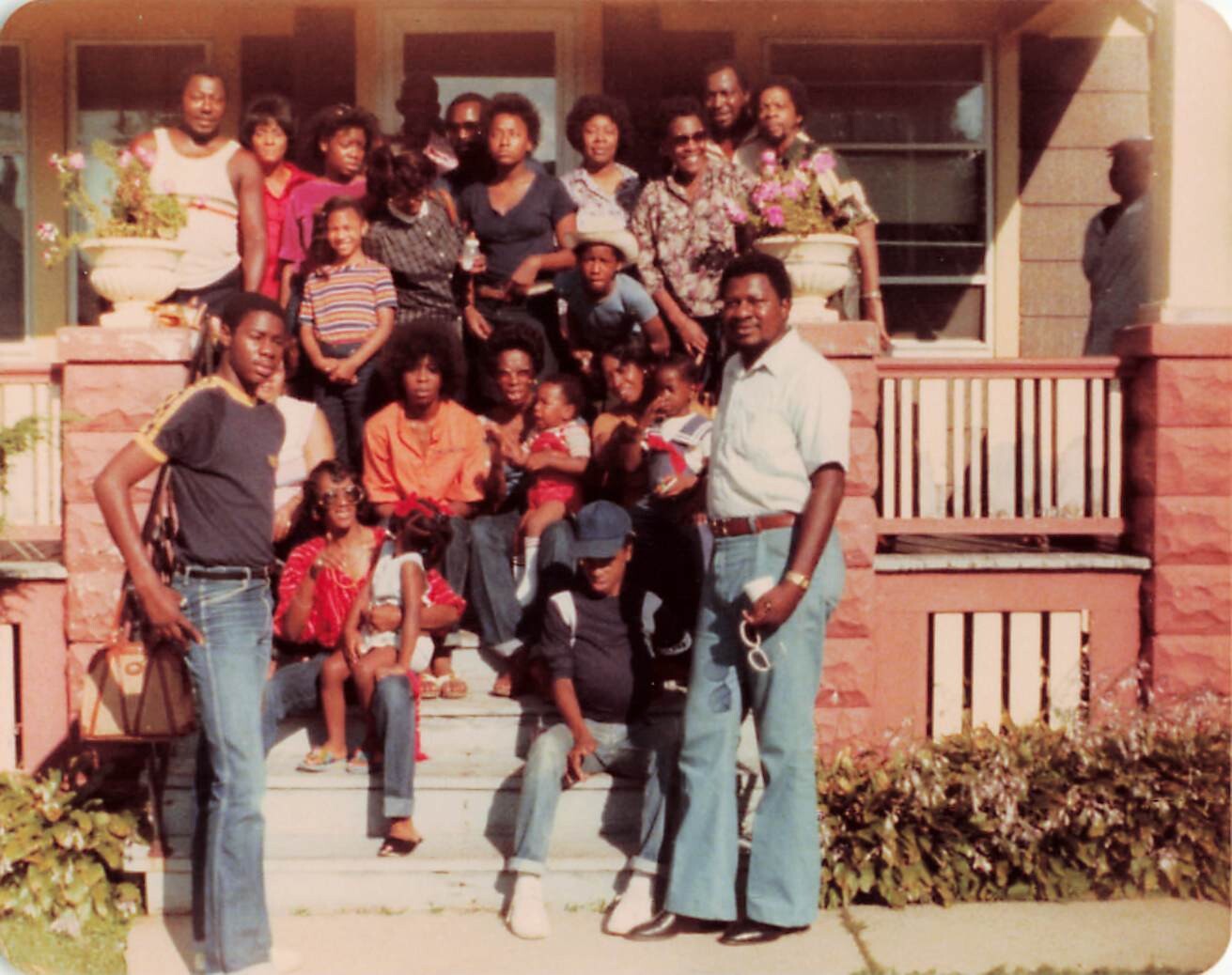 Large African American family posing on the front steps of a brick house, with adults, teens, and children gathered for a group photo on a sunny day.