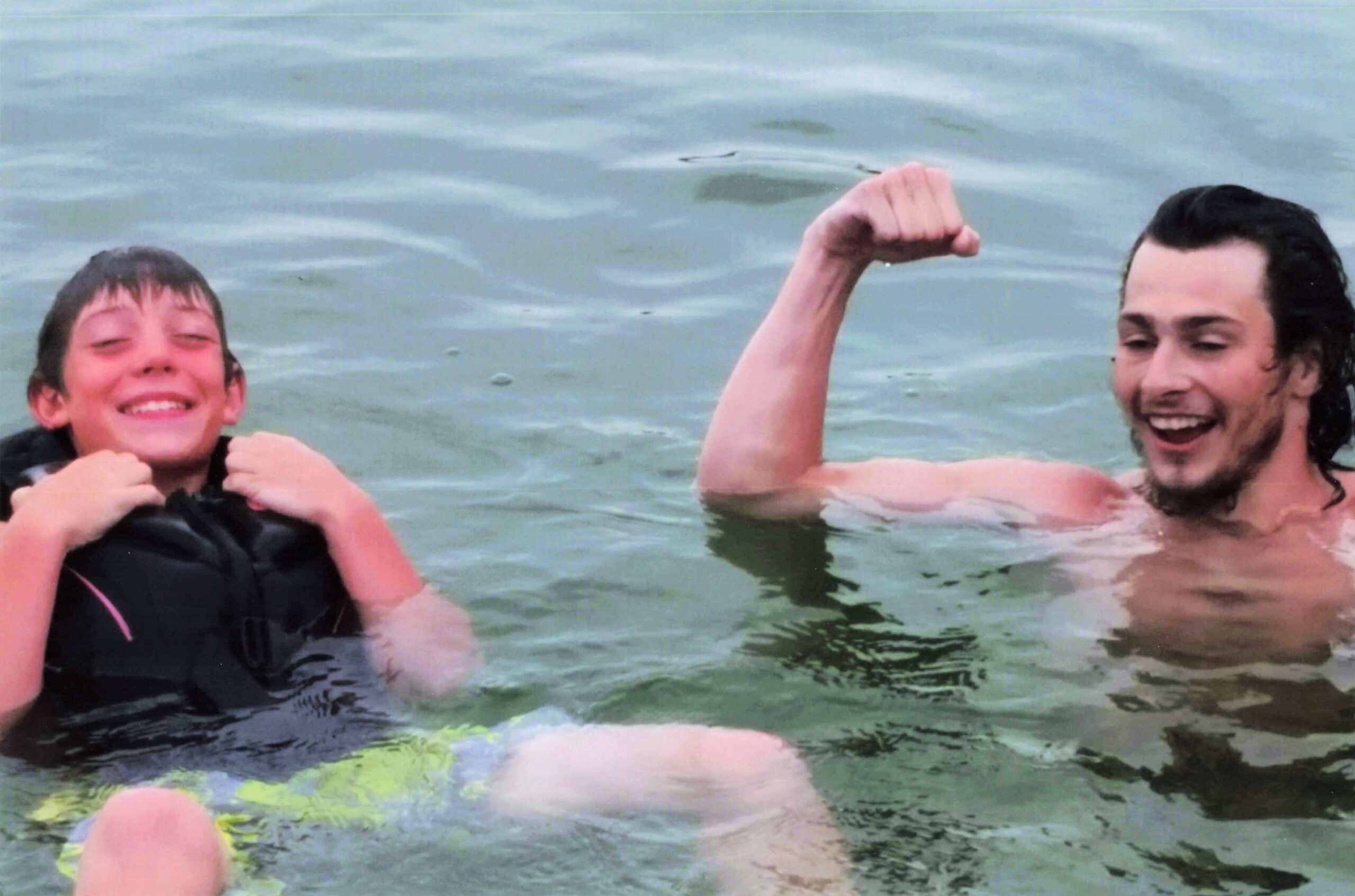 Two people swimming in a body of water, one smiling on the left and another on the right showing a flexed arm gesture.