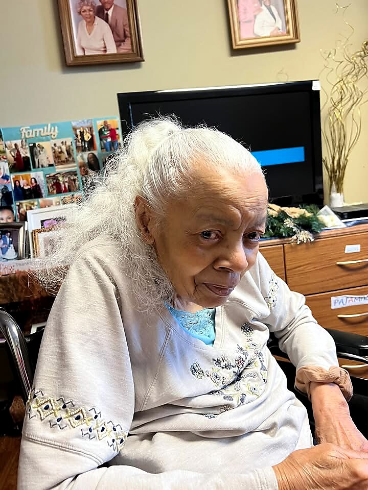 Older woman with white hair sits in a chair in a cozy living room, with a collage of family photos and a TV in the background.