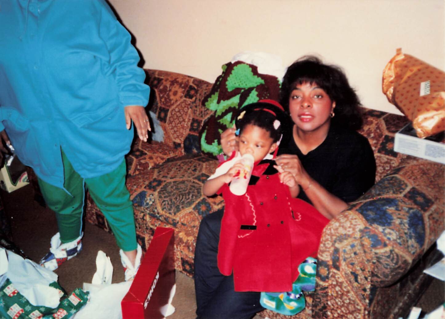 Young child in a bright red coat drinks from a bottle while a woman sits beside her on a patterned sofa amid gifts.