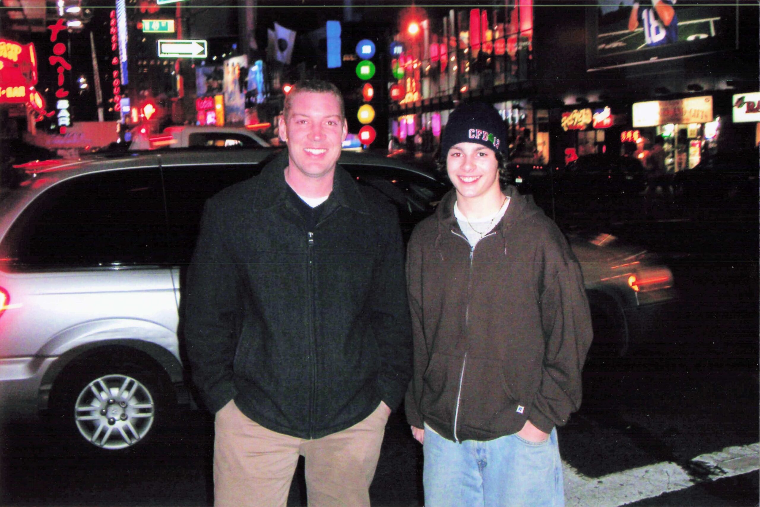 Two young men stand in front of a dark car on a busy neon-lit city street at night, smiling at the camera.